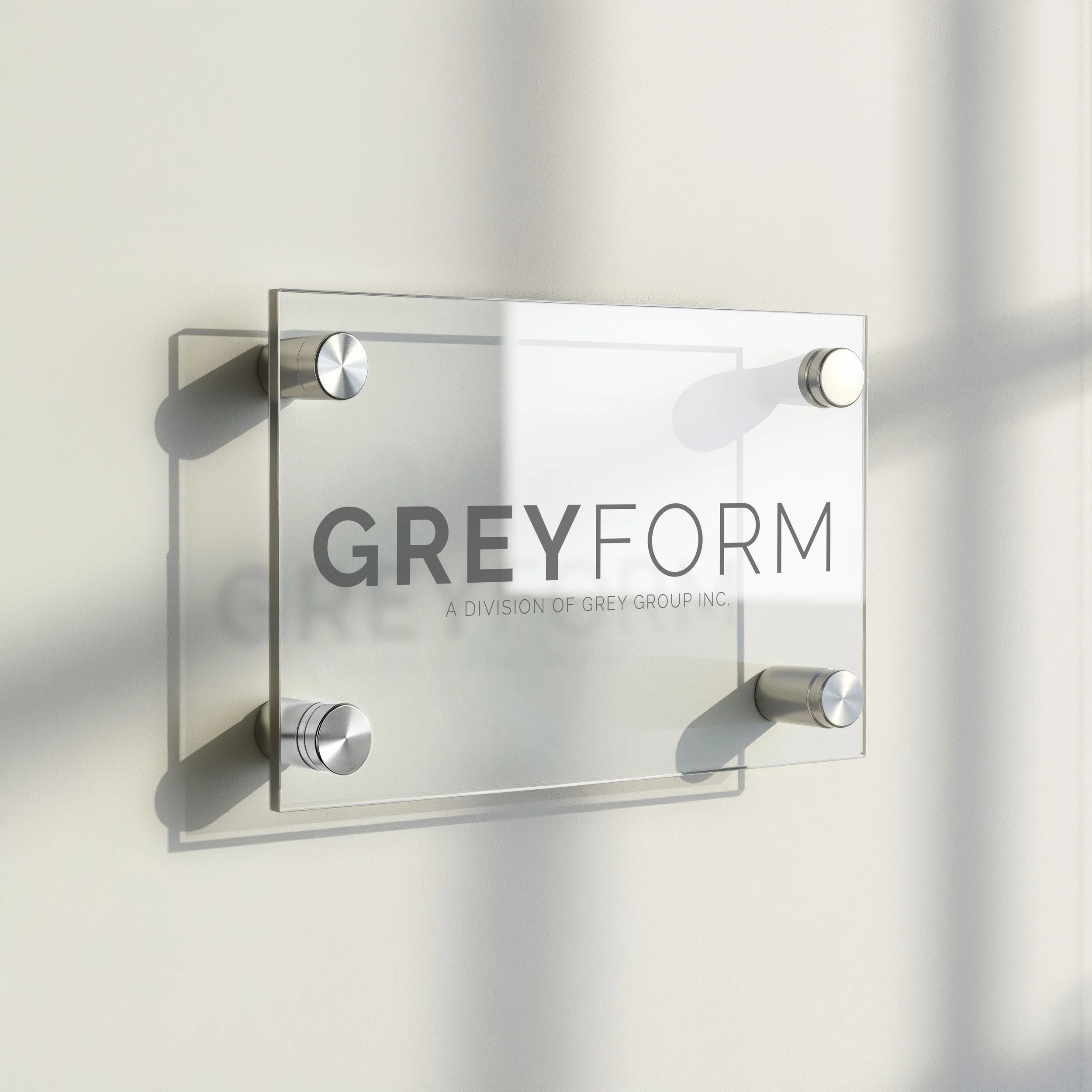 Modern clear glass sign with metal fasteners displaying the words 'GREYFORM' and 'A Division of Grey Group Inc.' mounted on a light-colored wall.