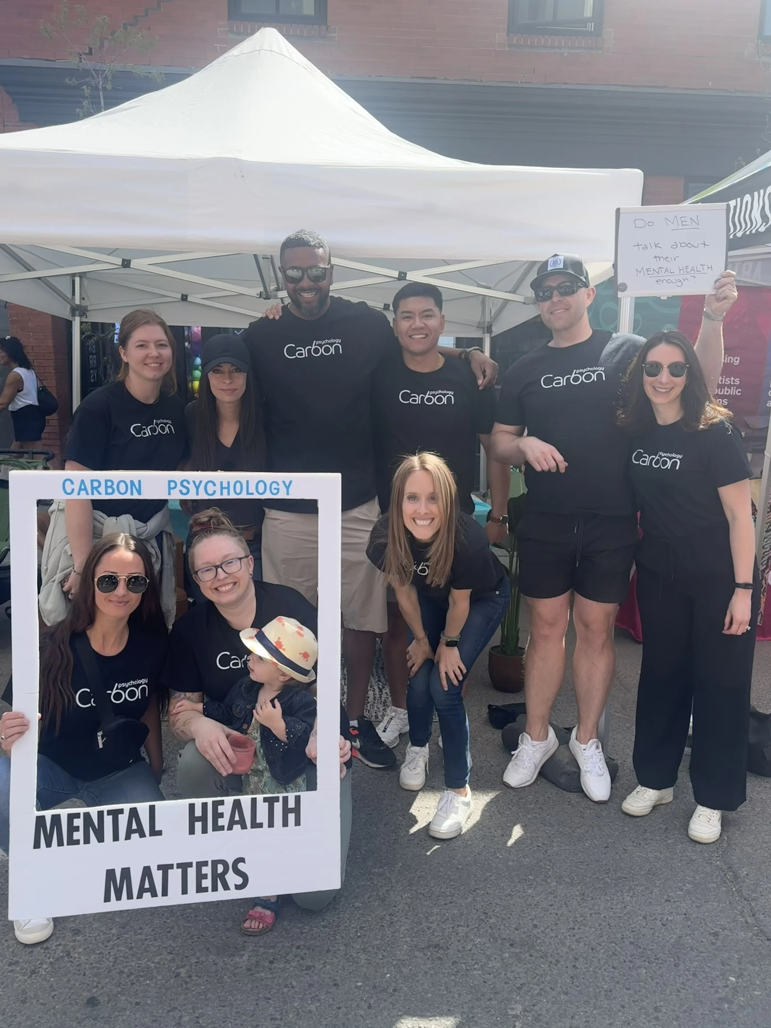 Group of six people standing under a white canopy tent at an outdoor event, wearing matching black t-shirts with 'psychology Carbon' printed on them, smiling, with water bottles and a blue sign that says 'psychology Carbon' in the background.