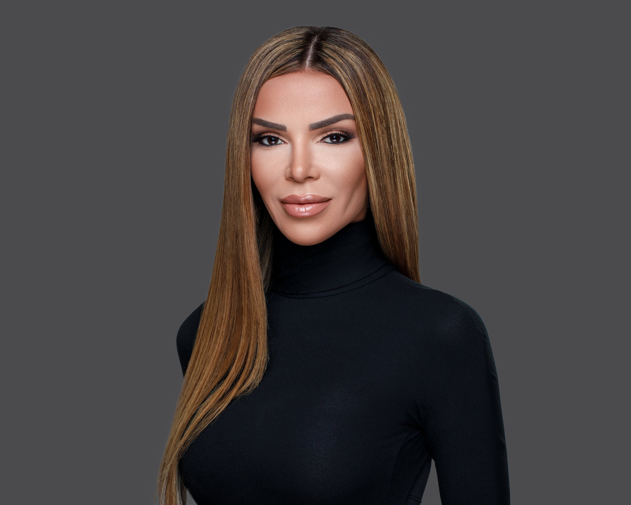 A woman with long straight hair wearing a black turtleneck posing against a gray background.