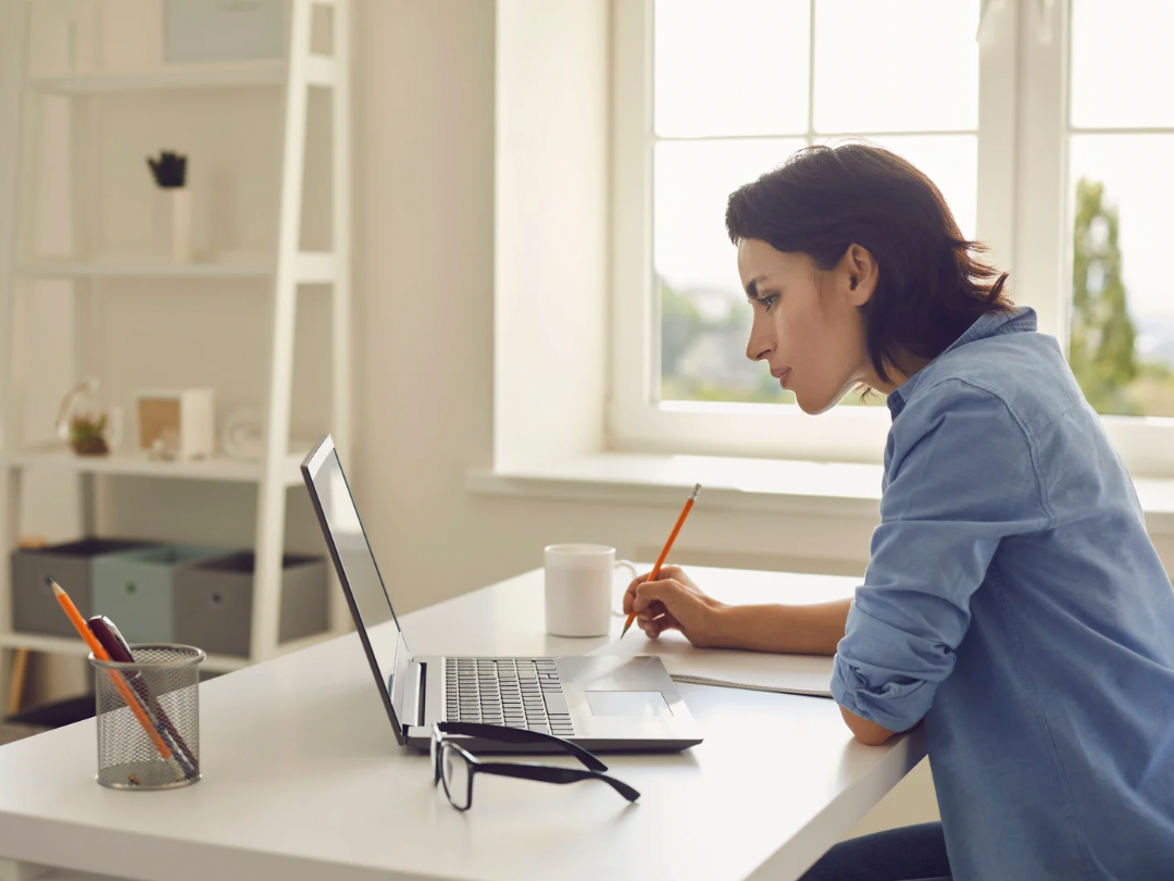 Woman working at a desk with a laptop, notebook, mug, and glasses in a bright room with large window.