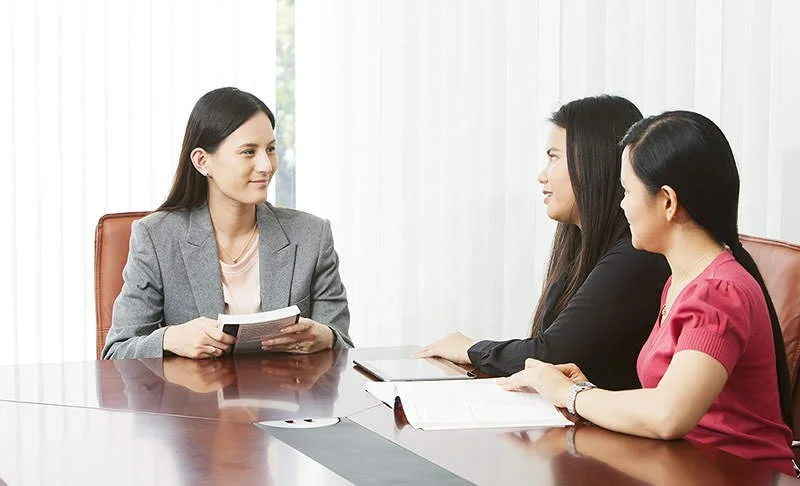 Jane Elderkin sitting in a boardroom holding a textbook, engaged in a discussion with two staff members about assessment design and teaching practices.