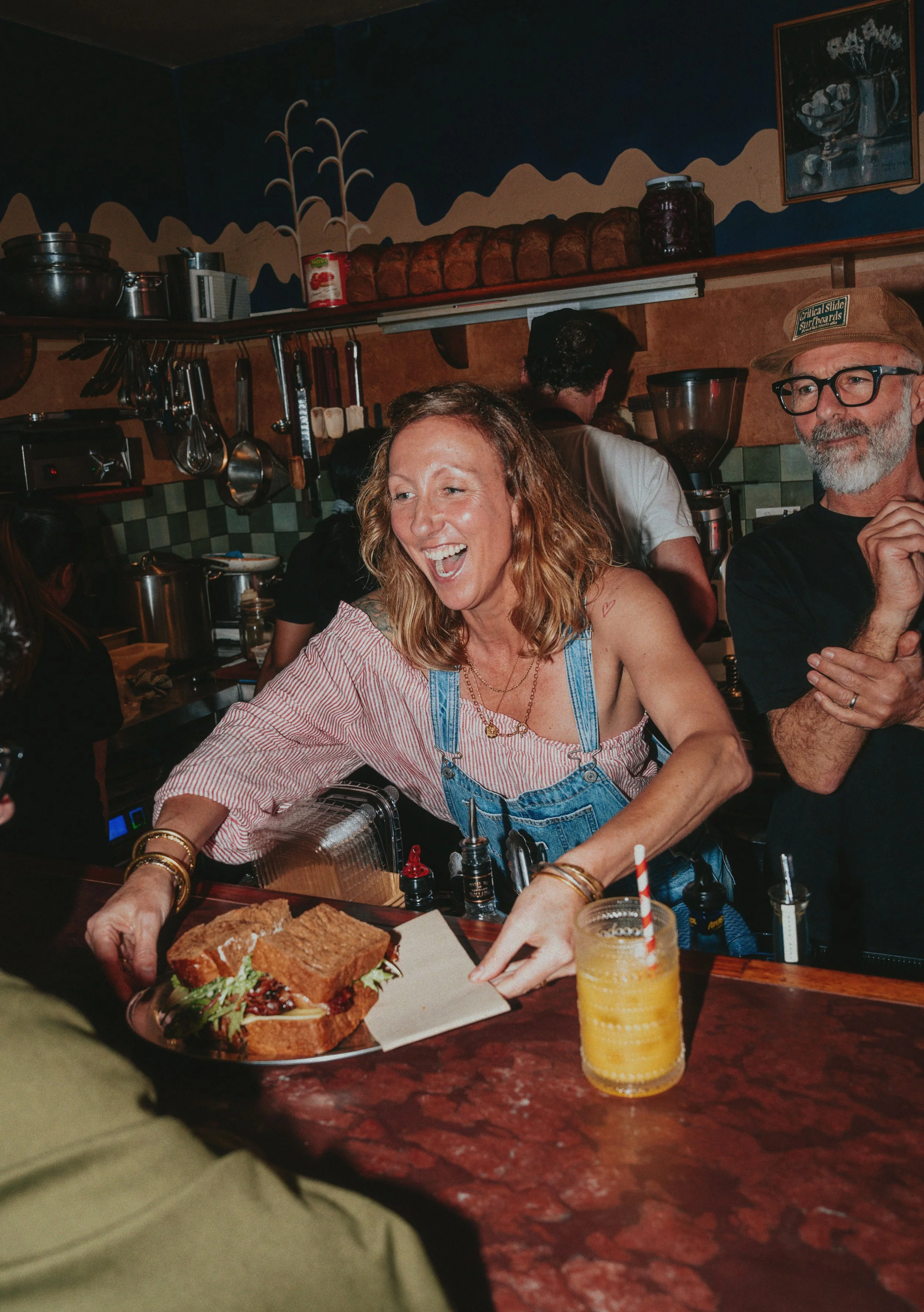 People enjoying a meal at a restaurant, with a woman smiling and a man wearing glasses and a cap. There's a sandwich, a yellow drink, and condiments on the table.