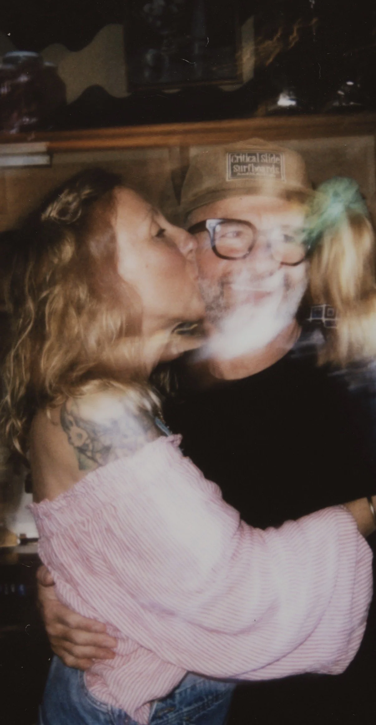 A woman with long wavy hair kissing an older man wearing glasses and a hat, both smiling and embracing each other.