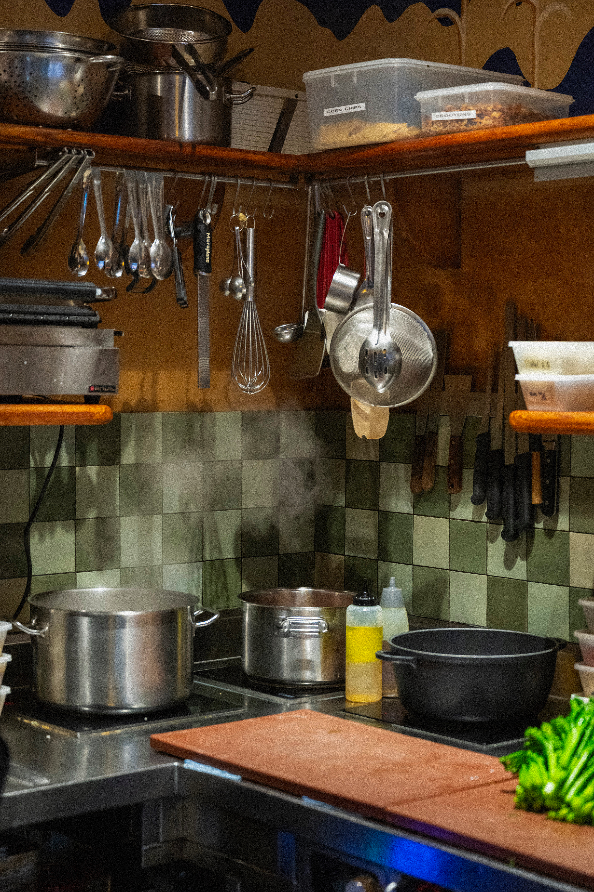Kitchen scene with pots on the stove, hanging utensils (ladle, whisk, measuring cups), a plastic container with labeled compartments containing corn chips and croutons, and a cutting board with lettuce. The background features a tiled wall and wooden