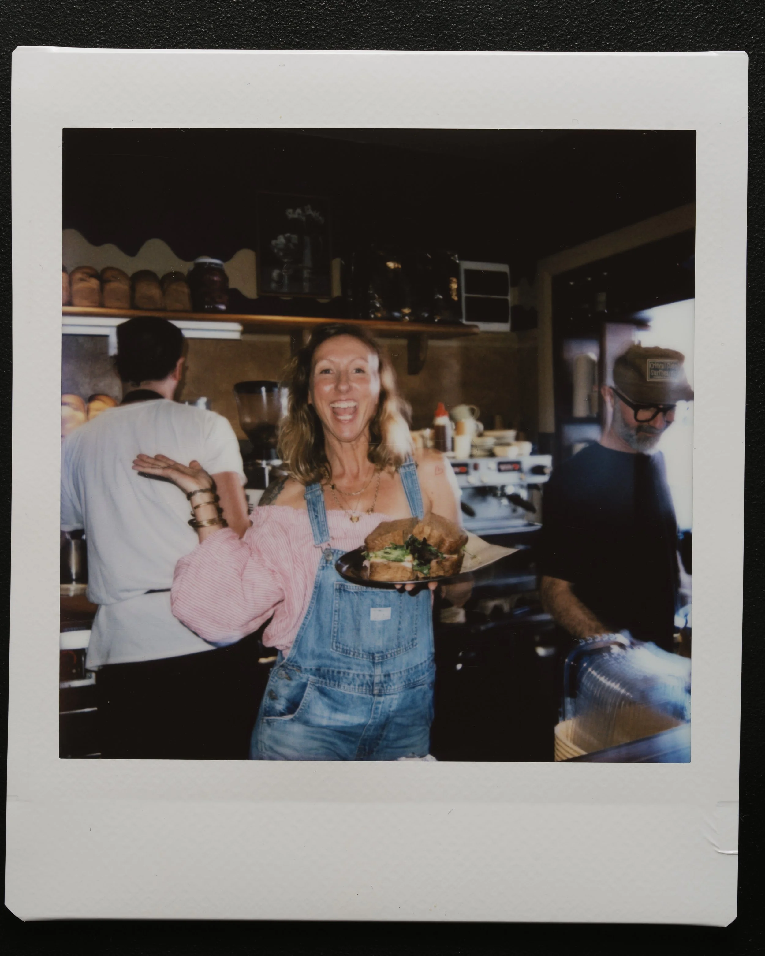 A woman in denim overalls and pink shirt holding a plate of food with toast, greens, and an omelet, smiling in a kitchen with a woman in white with short dark hair and a man with glasses.