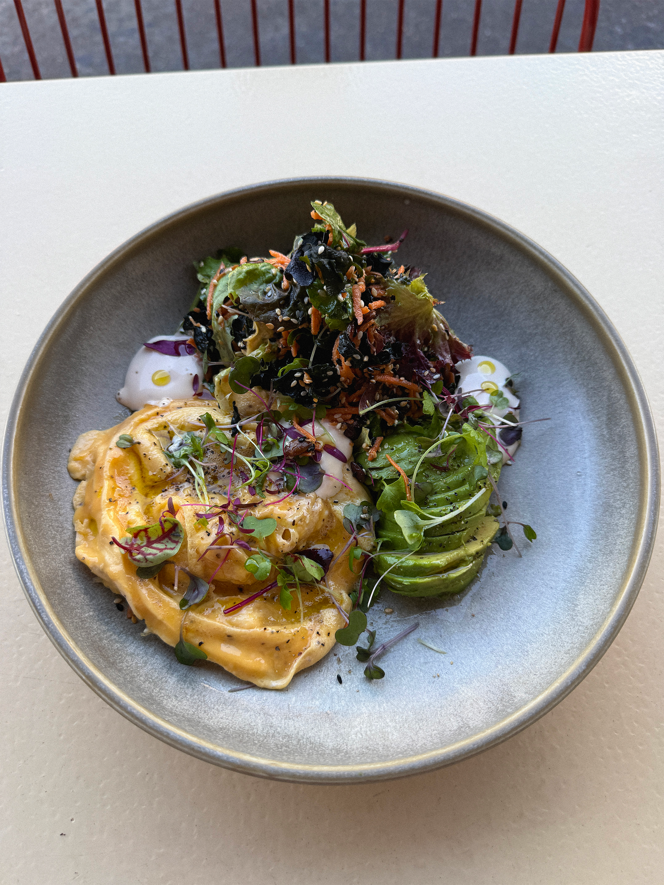 A plate of food with a fried or baked egg topped with microgreens, alongside a colorful salad with various greens, shredded carrots, and black sesame seeds, served on a gray ceramic plate.