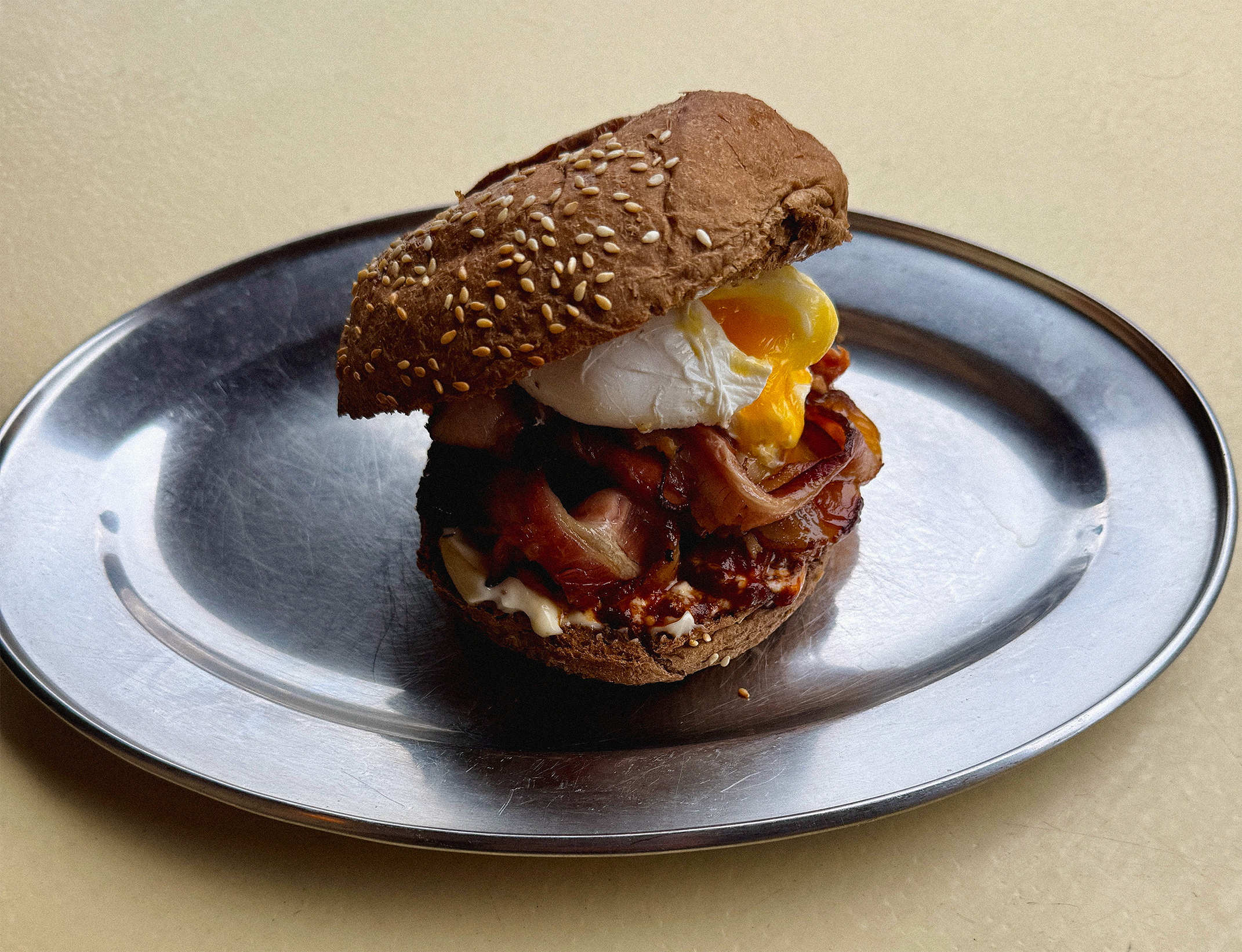 Close-up of a breakfast sandwich with a sesame seed bun, fried egg, bacon, and melted cheese on a silver tray.