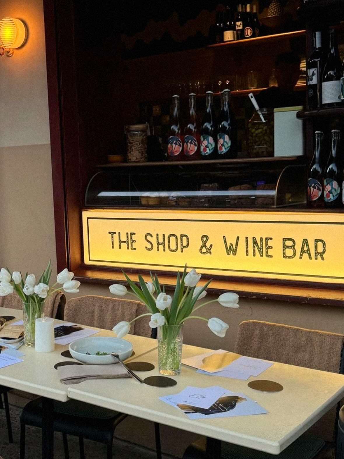 A table set with white flowers in vases, plates, napkins, and menus, in front of a sign that reads 'The Shop & Wine Bar'. Behind the table, there is a dark wooden counter with bottles and glasses, and a hanging light fixture on the wall.