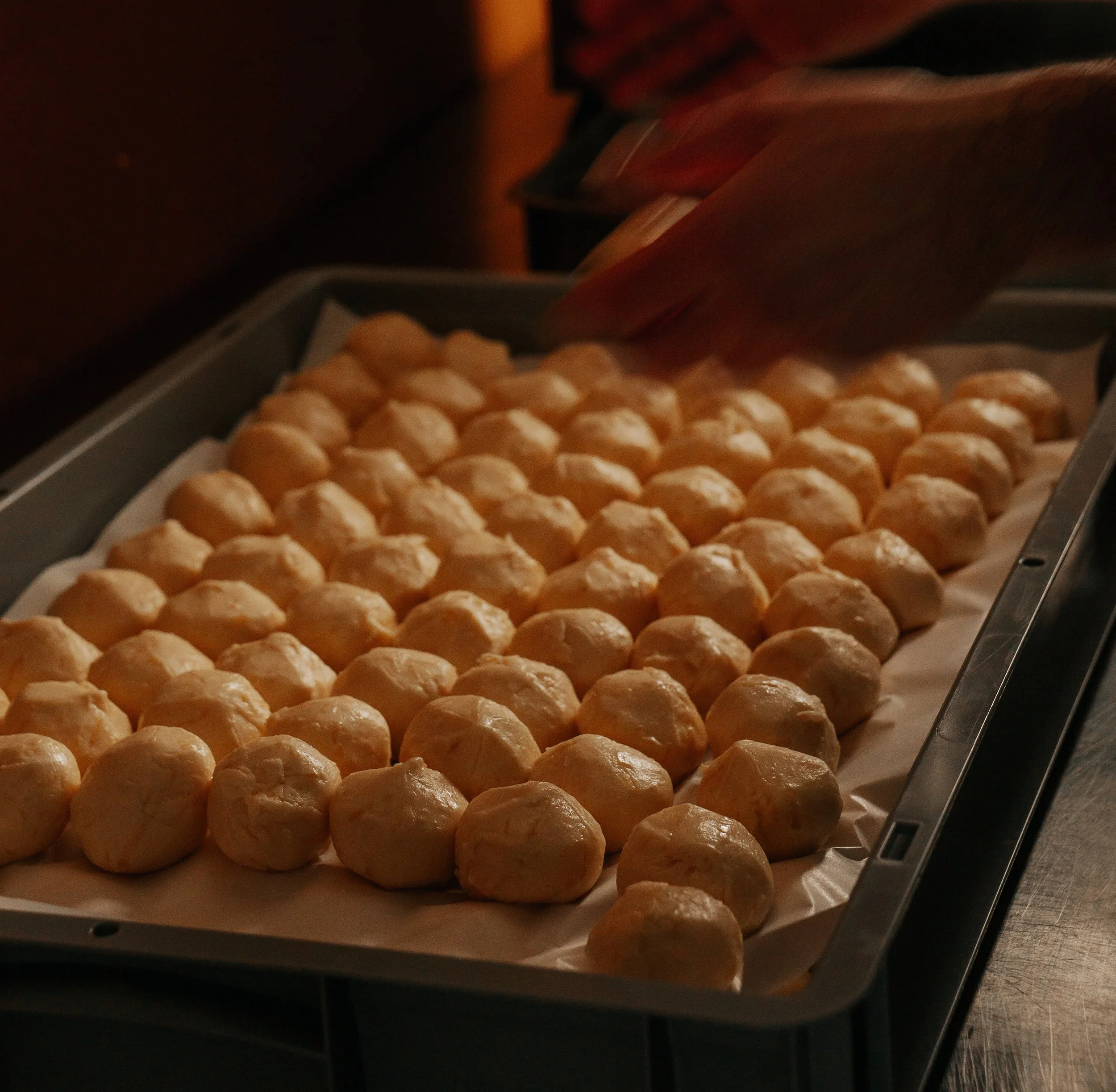 Tray of freshly rolled pão de queijo in Lova's kitchen with a hand rolling and placing a new ball of dough on the tray.