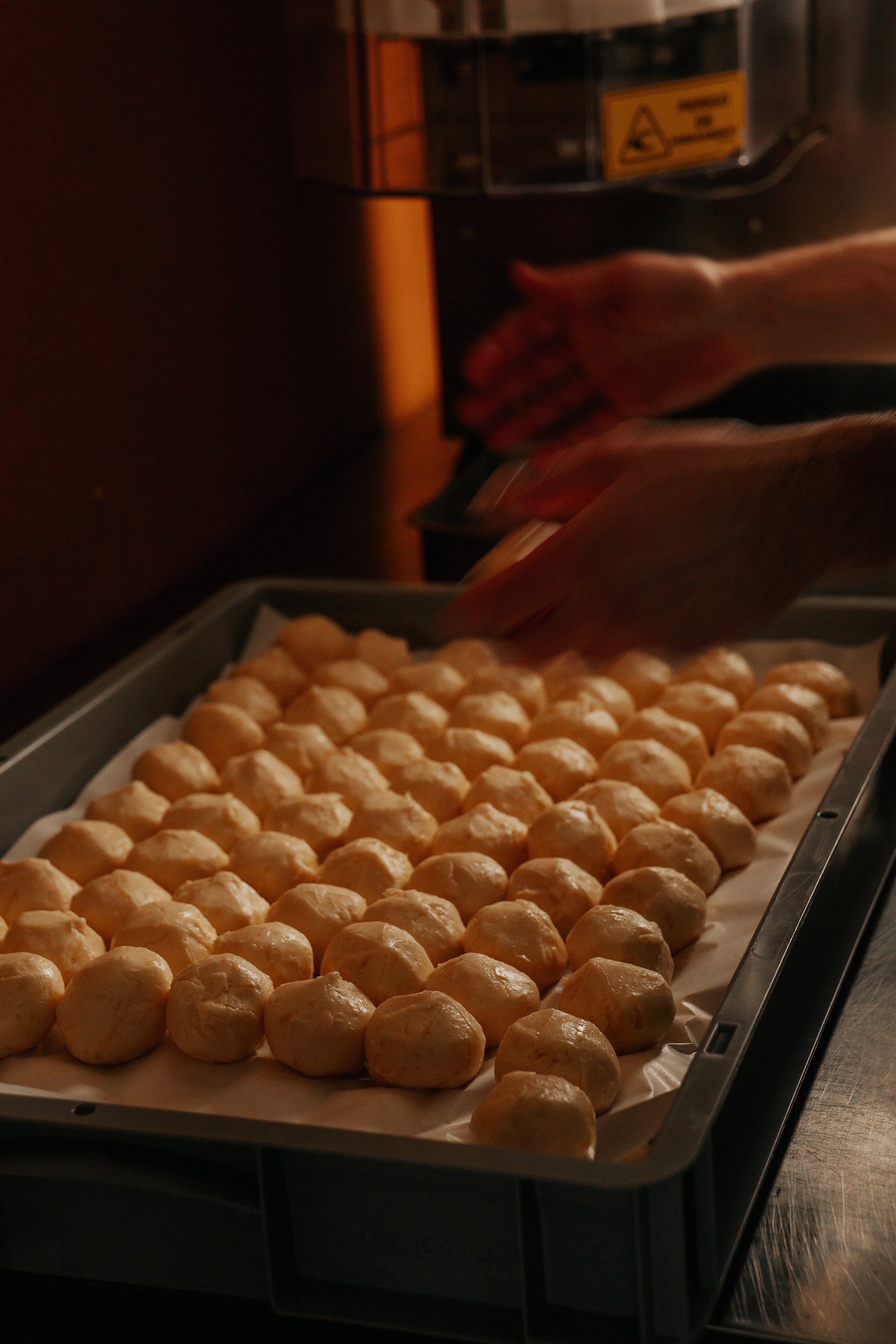 Tray of freshly rolled pão de queijo in Lova's kitchen with a hand rolling and placing a new ball of dough on the tray.
