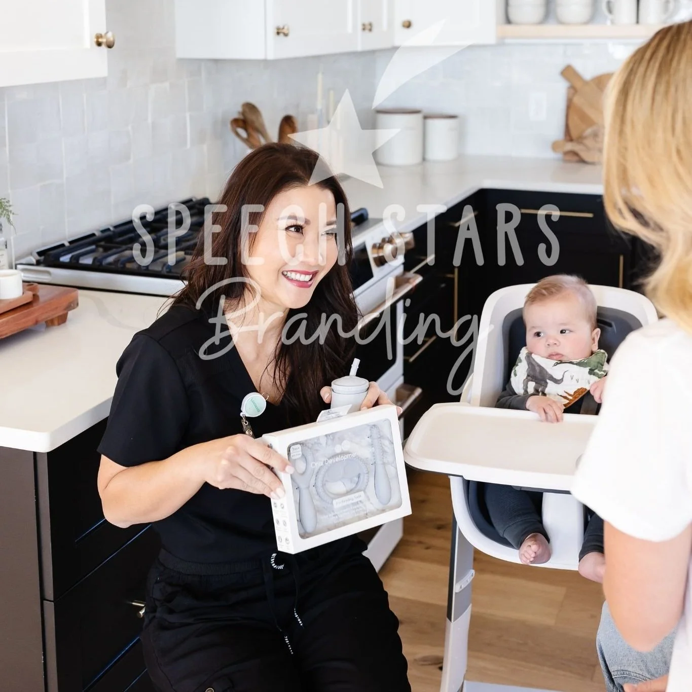 Woman with dark hair smiling and holding a baby care item, sitting in a kitchen near a baby in a high chair and another woman standing nearby.