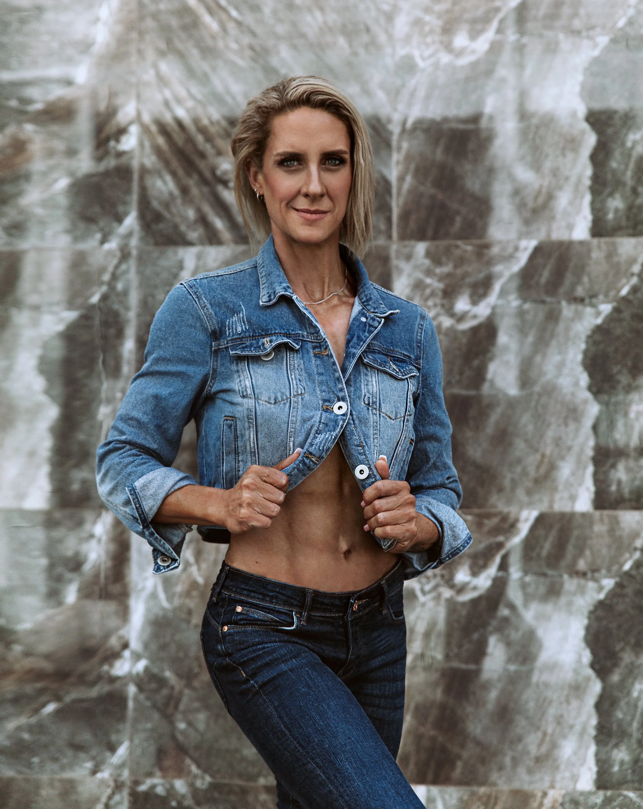 A woman with short blonde hair wearing a denim jacket and jeans standing in front of a marble wall.