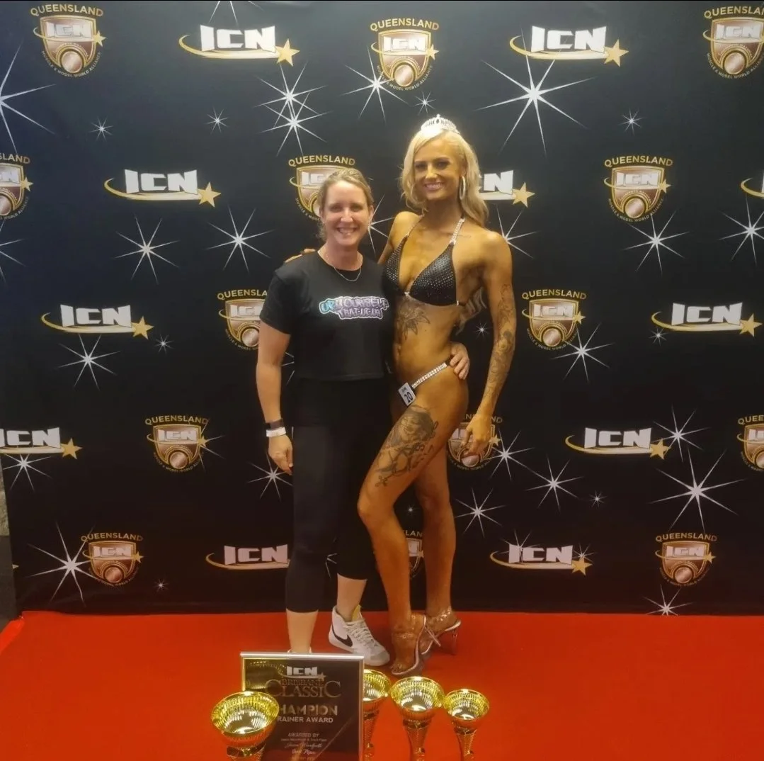 Two women standing in front of a backdrop with logos, one in sports attire and the other in a swimsuit and high heels, celebrating an event with trophies and a certificate.