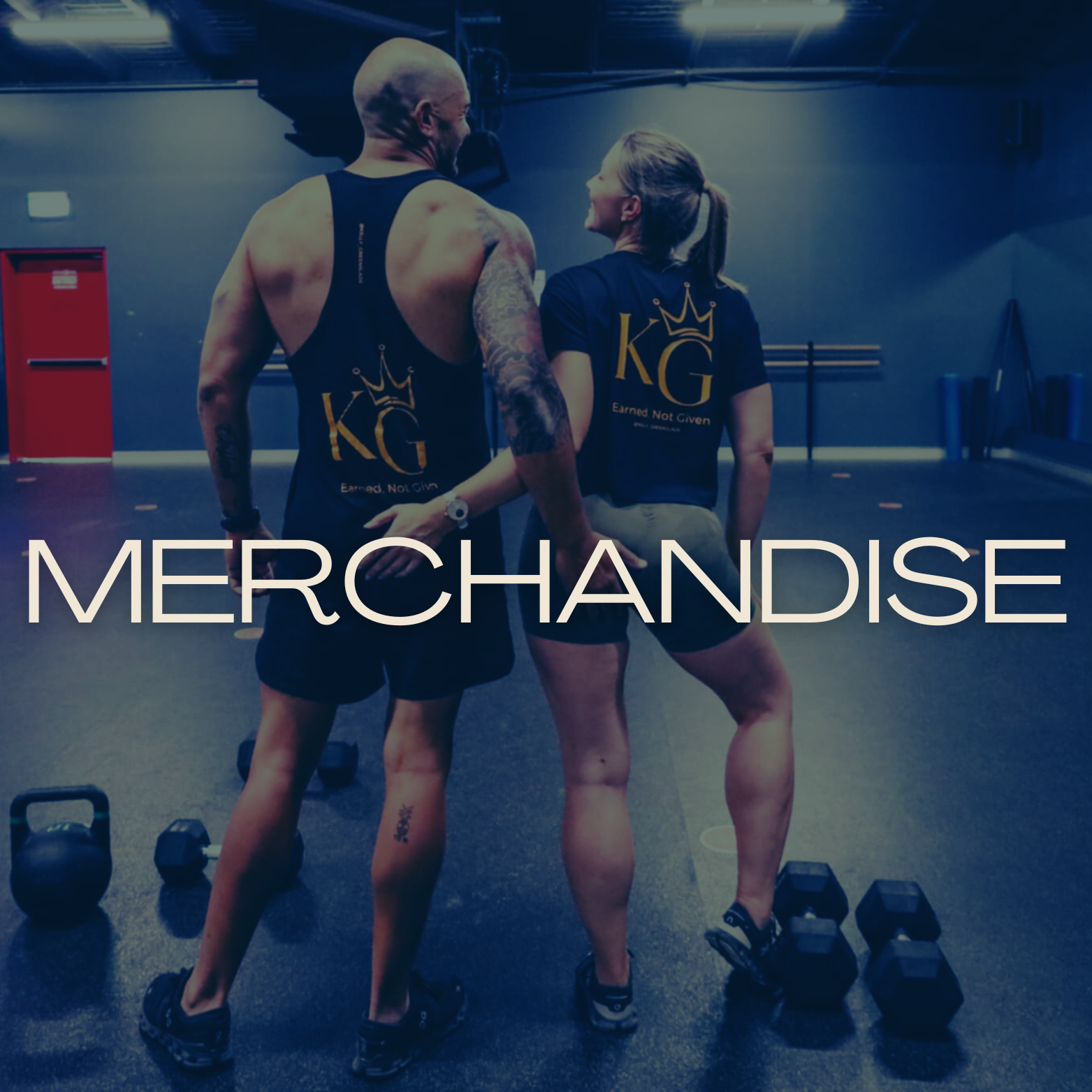 Two people stand in a gym, smiling and holding hands, dressed in matching black and yellow workout shirts that say 'KG' with a crown and 'Earned Not Given' underneath. The gym has dumbbells on the floor and workout equipment in the background. The word 'MERCHANDISE' is overlaid on the image.