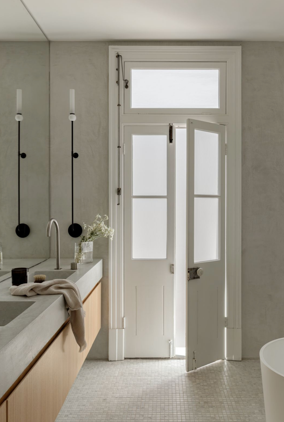 Black bathroom wall lights beside original French doors with frosted glass and a transom window, above a timber vanity with a grey basin top in a microcement bathroom.