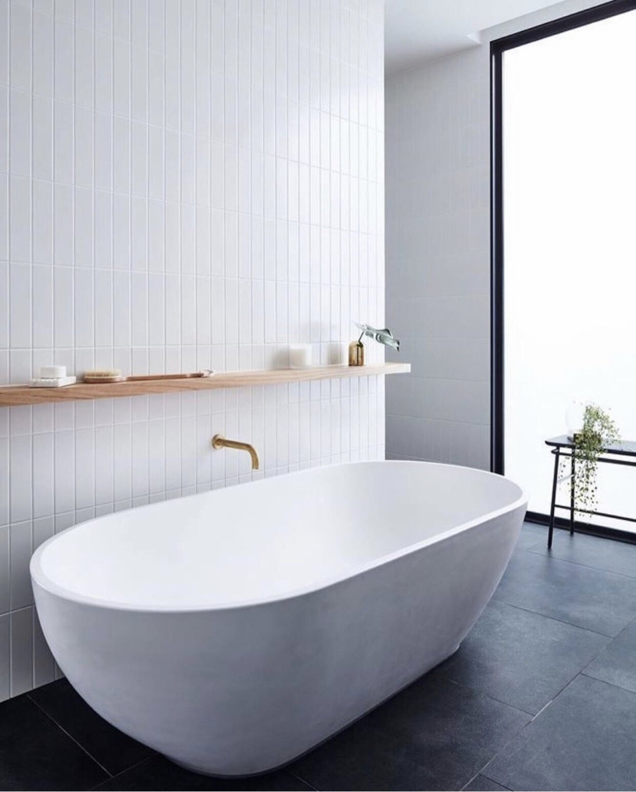 Stacked tile in a vertically stacked tiling pattern in a modern bathroom with gray floor tiles and a white freestanding bath and wooden bath shelf