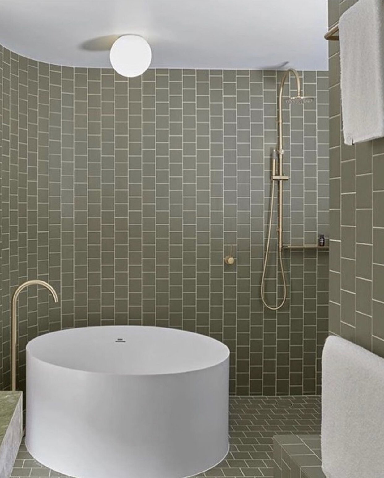 green square tiles glued to brick on bathroom floors and walls, curved wall behind bath and brass shower head and tapware