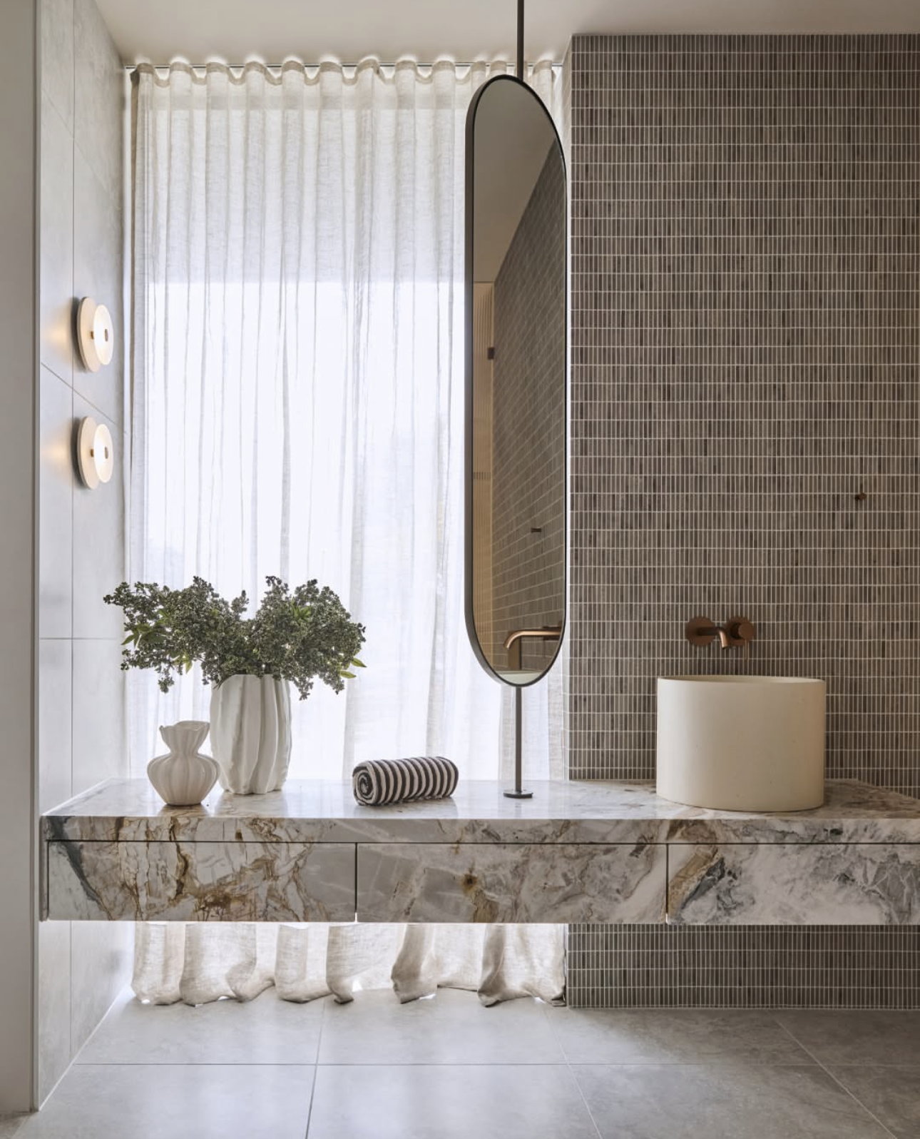 Floating marble bathroom vanity with wall lights, hanging oval mirror and natural light from a sheer-curtained window