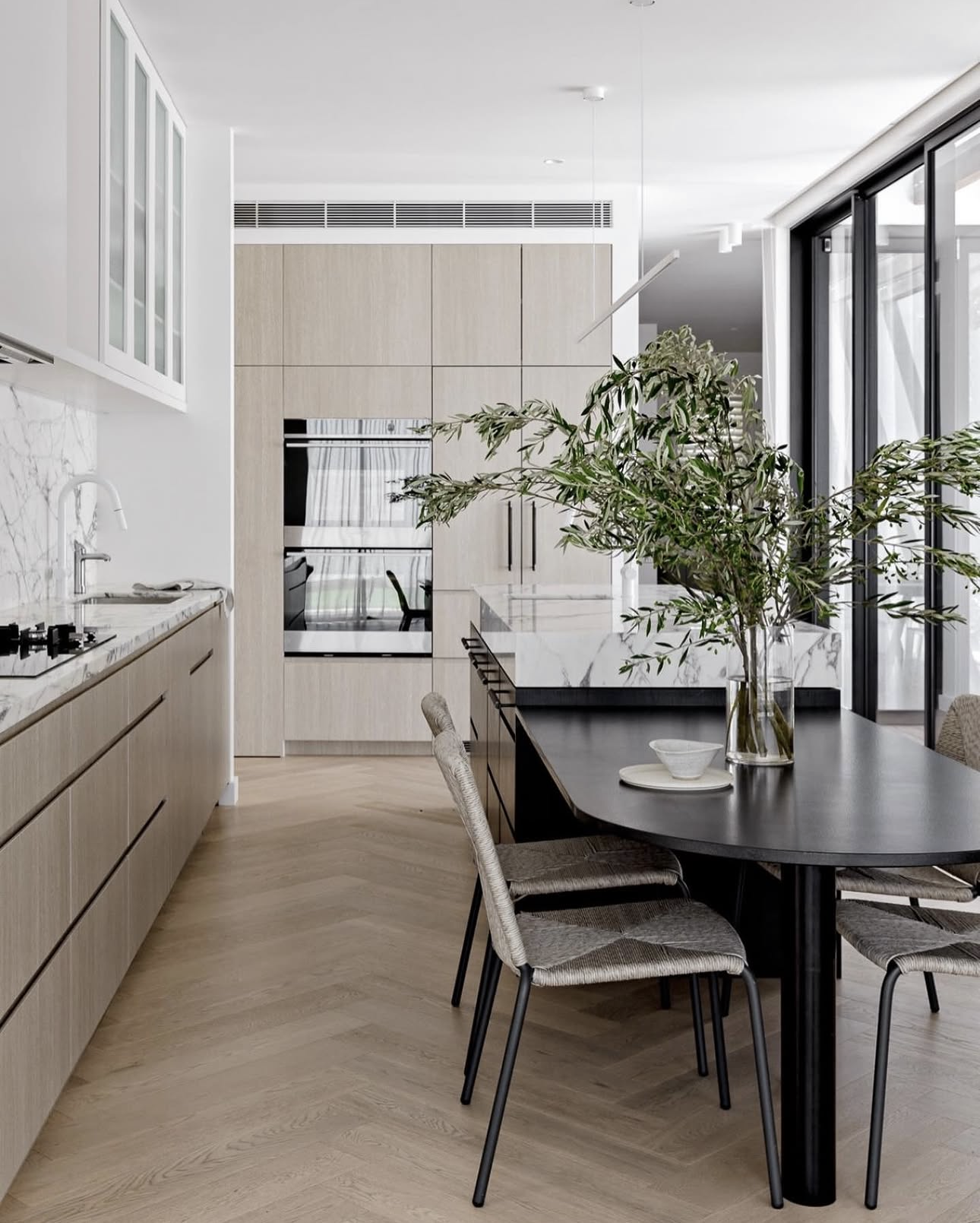 Black oval dining table attached to the end of a kitchen island bench, with upholstered dining chairs