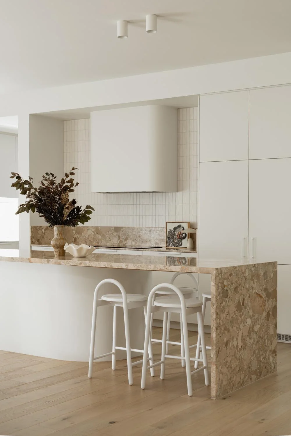 Stone kitchen island bench at bench height with white bar stool seating at end and along the back