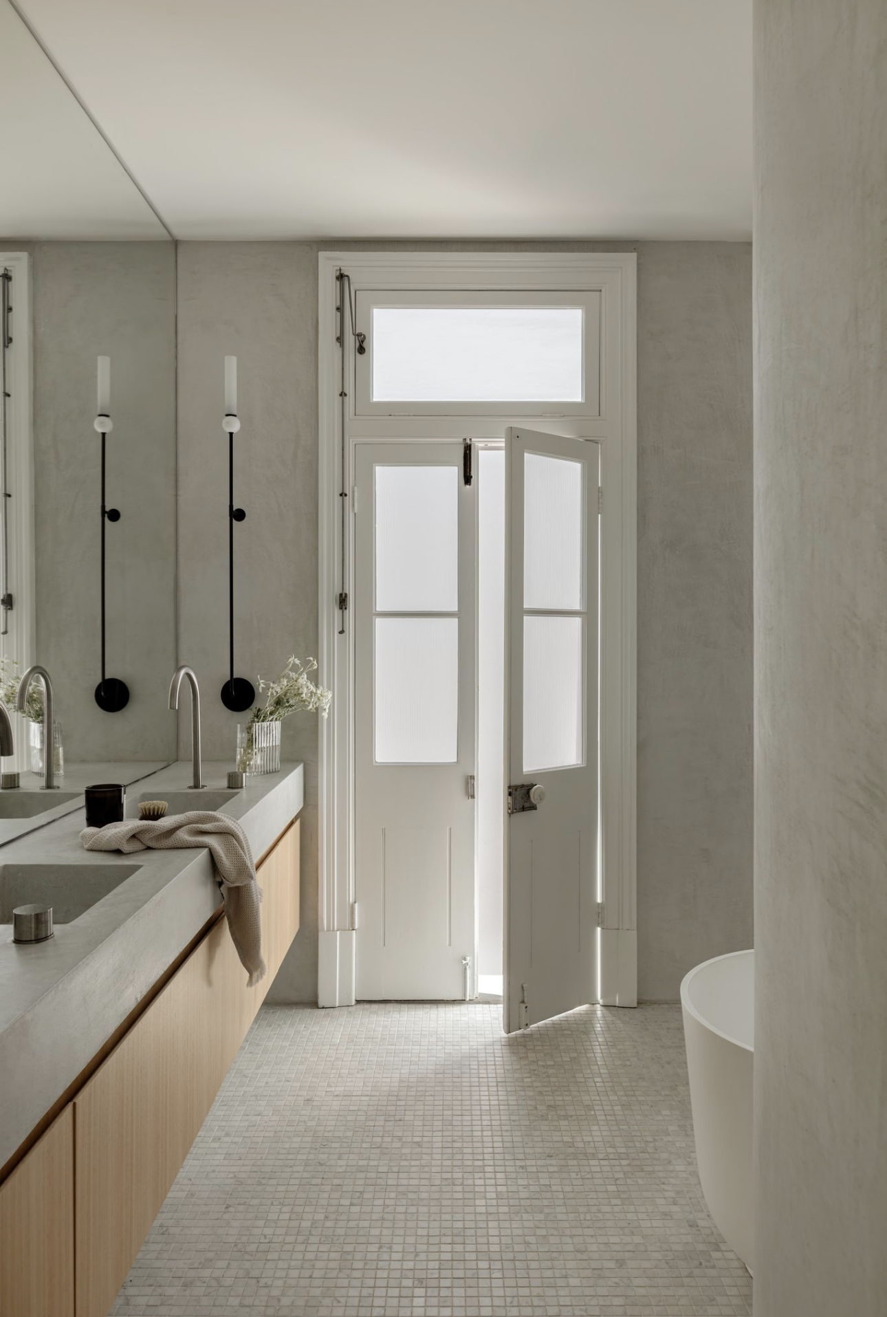 Microcement bathroom with original French doors, a freestanding bath and a double timber vanity on a mosaic tiled floor.