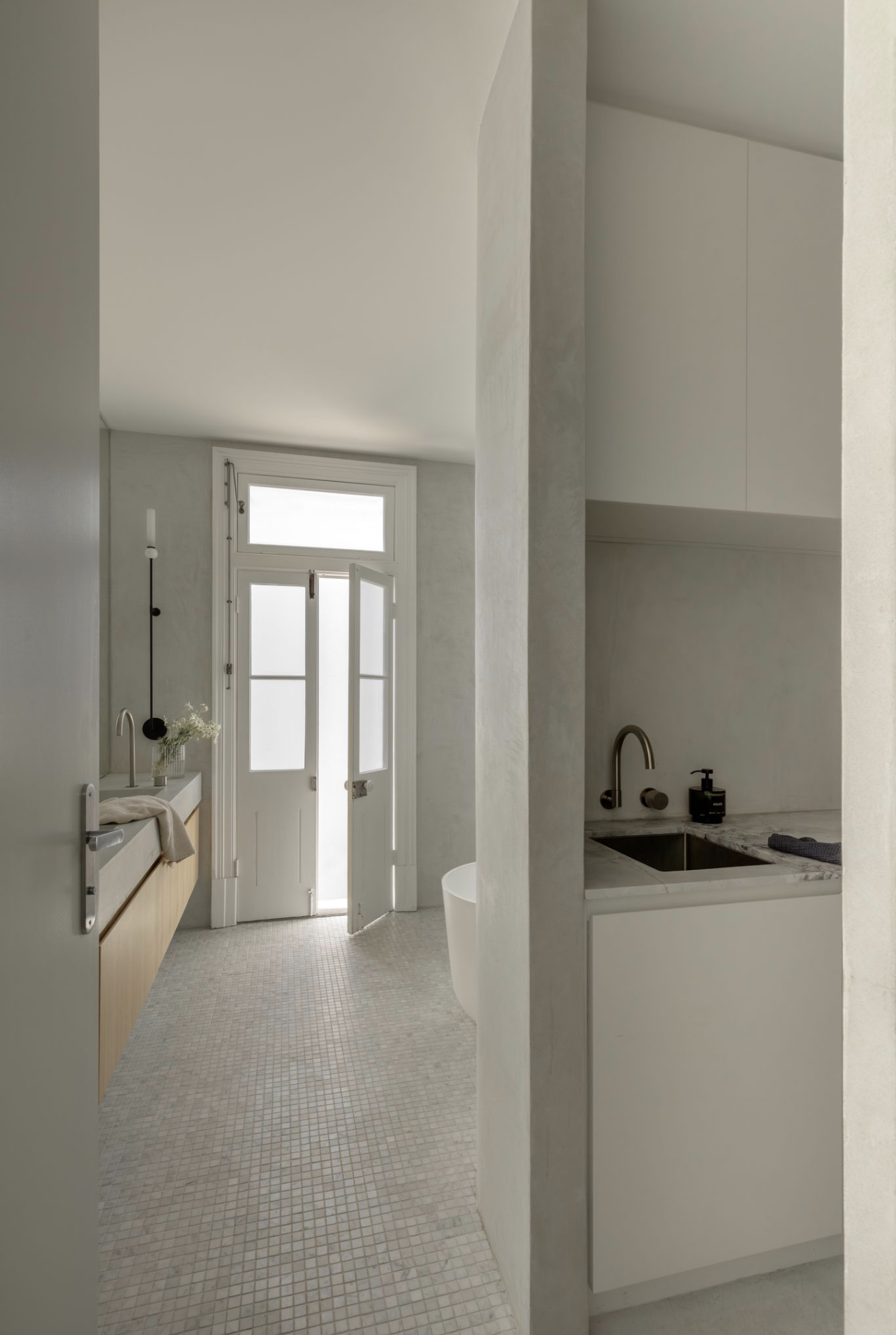 Bathroom laundry combo with a sink and cabinetry tucked behind a curved microcement wall, looking through to the original French doors, a freestanding bath and a timber vanity.