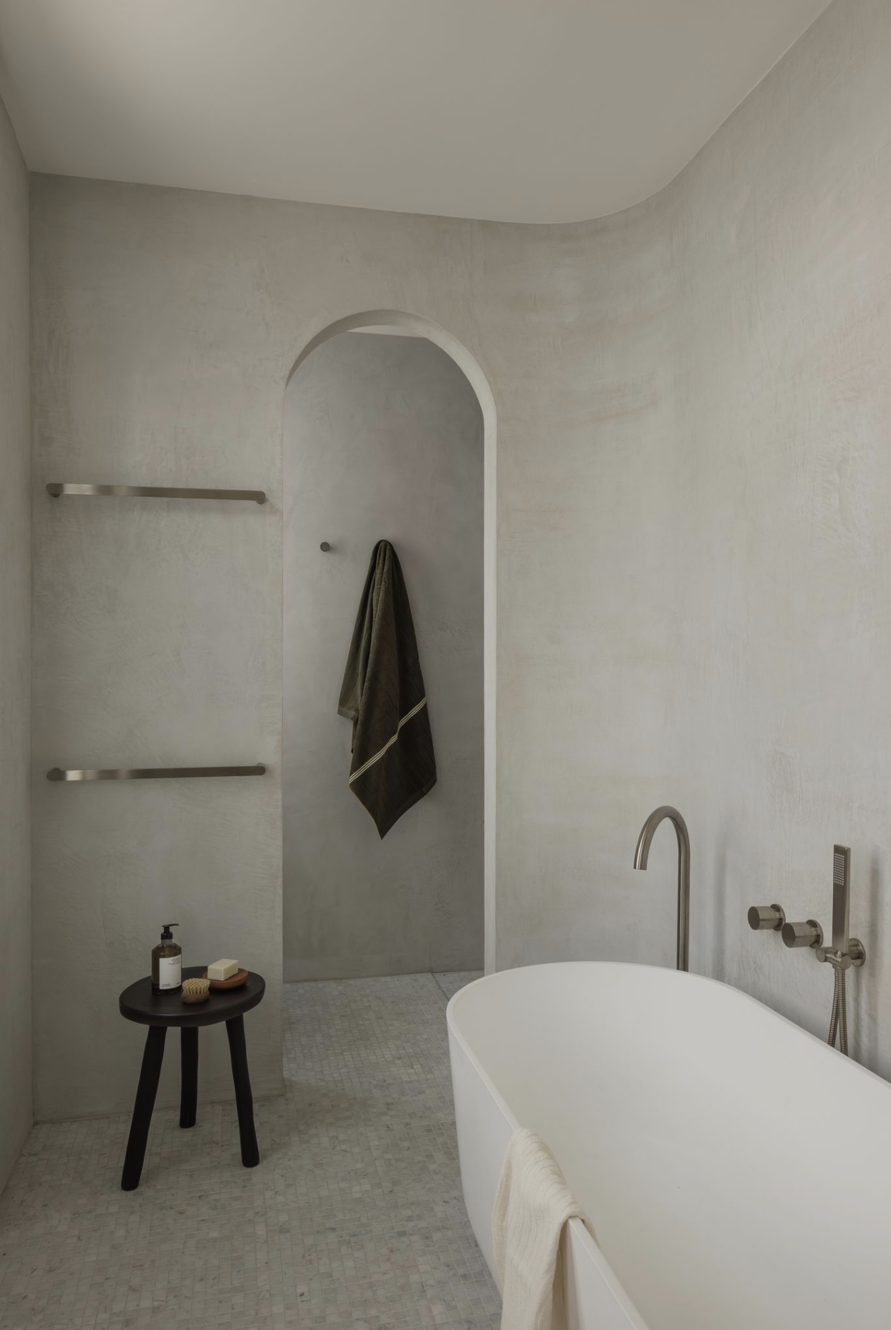 Arched opening in a microcement bathroom, with a freestanding bath, heated towel rails and a timber stool.