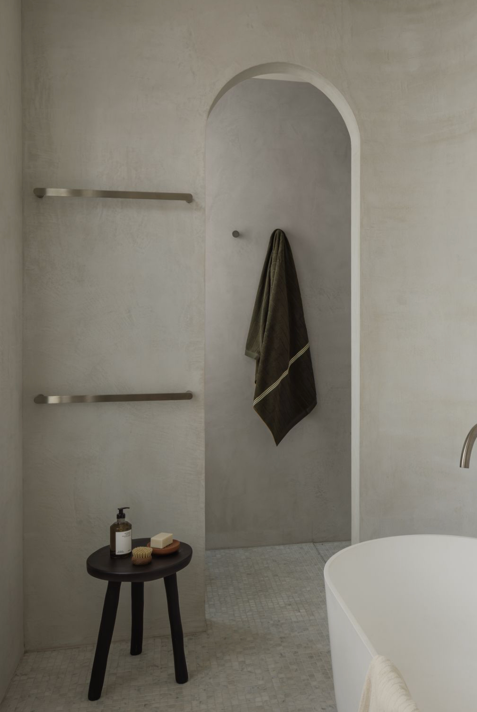 Arched doorway in a microcement bathroom, with towel rails on the wall, a dark timber stool and the edge of a freestanding bath on a mosaic tiled floor.