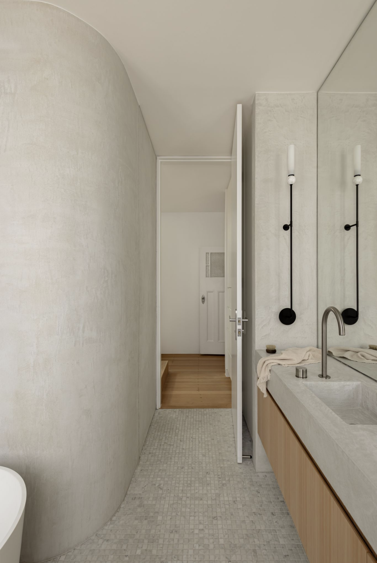 Curved microcement bathroom wall beside a timber vanity, with black wall lights and mosaic tiles on the floor.