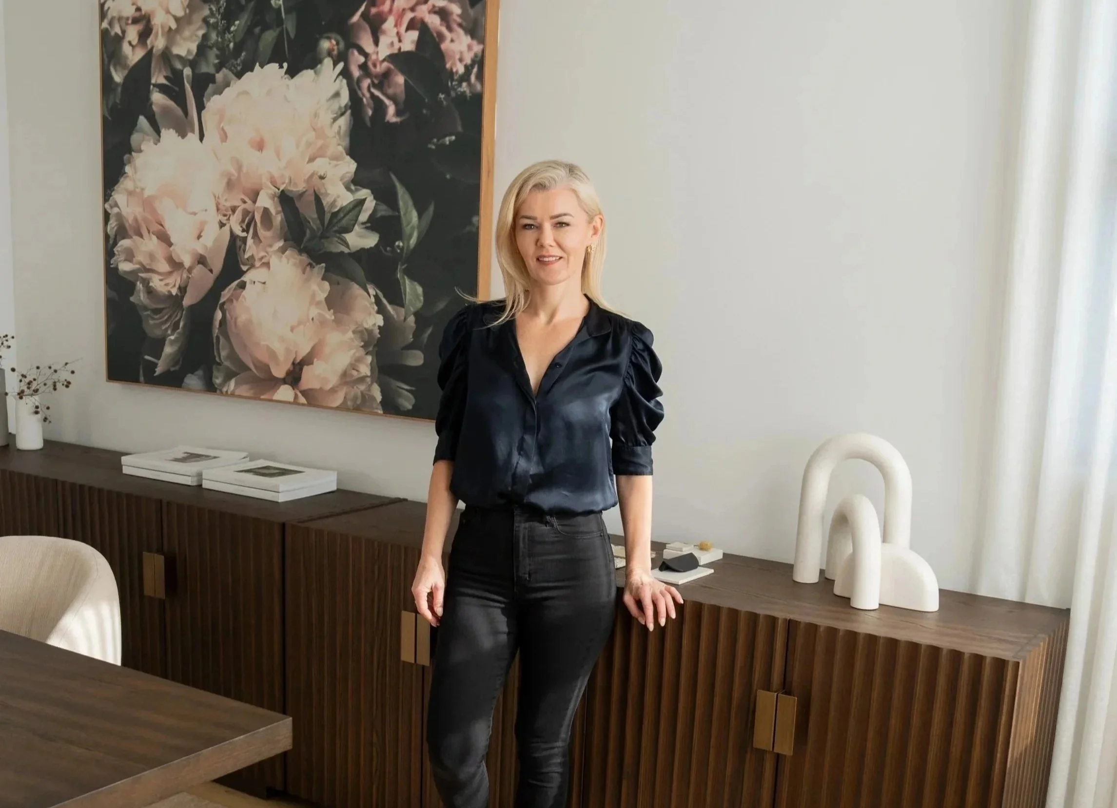A woman with blonde hair wearing a black top and black pants standing in a modern interior space with minimalist decor.