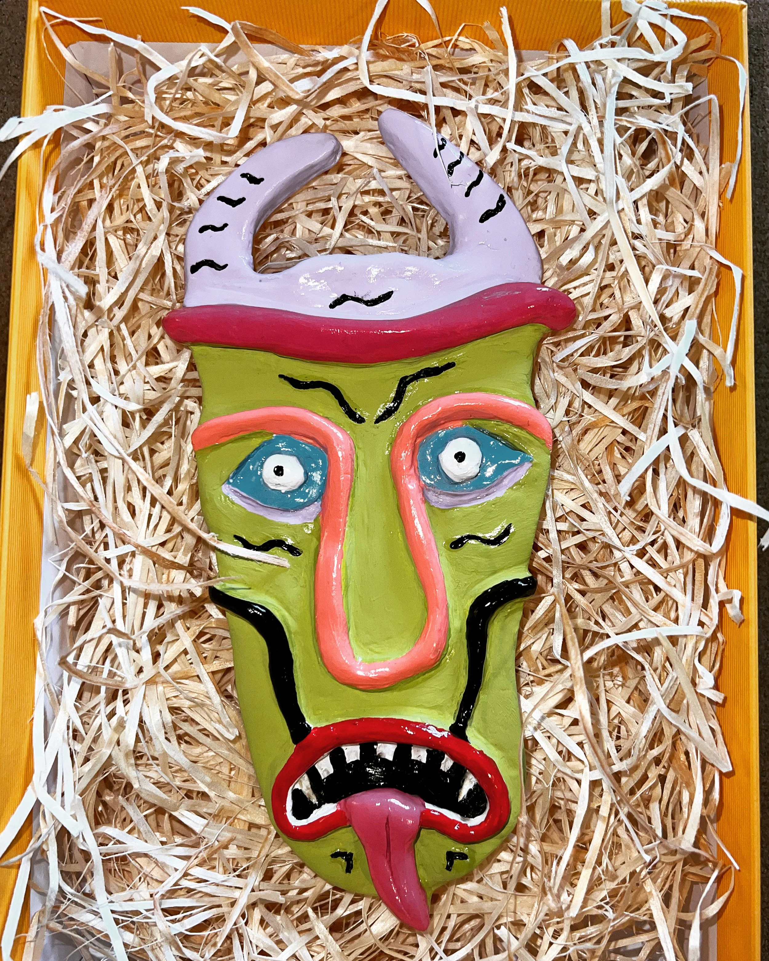 Colorful tribal-style mask with horns, large eyes, and a long tongue, placed on shredded paper inside a yellow box.