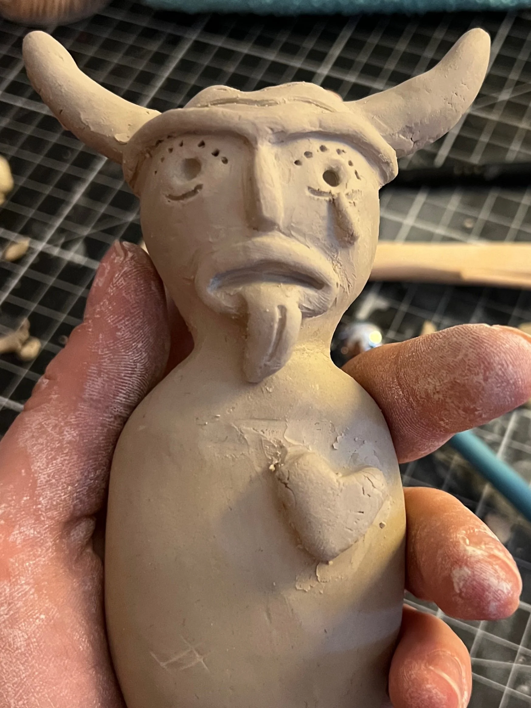A ceramic figurine of a mythical creature with a humanoid face, Viking-style helmet with horns, and a heart-shaped detail on its chest.
