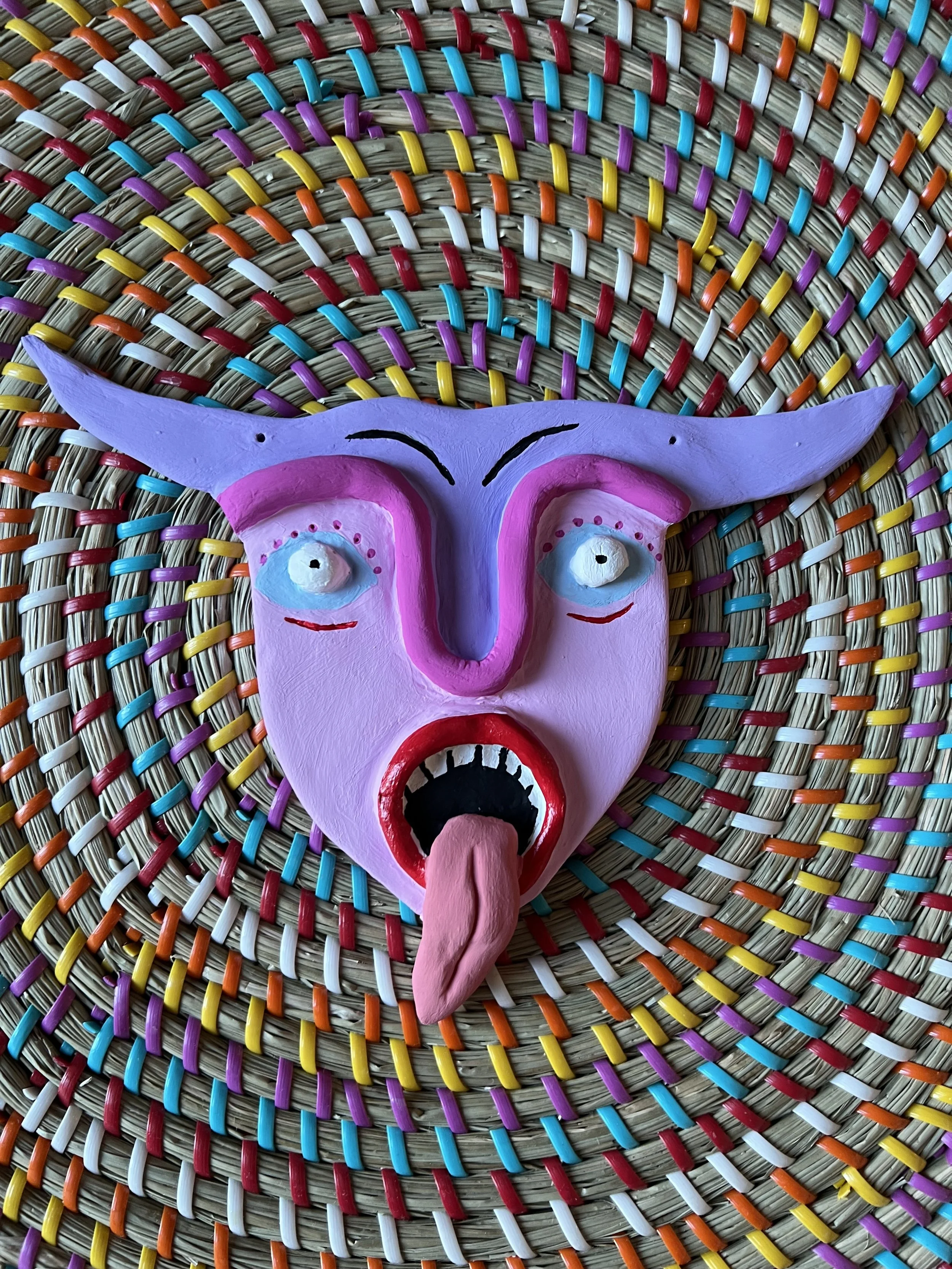 Colorful, stylized mask with a pink face, blue eyes, exaggerated eyebrows, purple horns, red lips, and sticking out tongue, on a multicolored woven background.