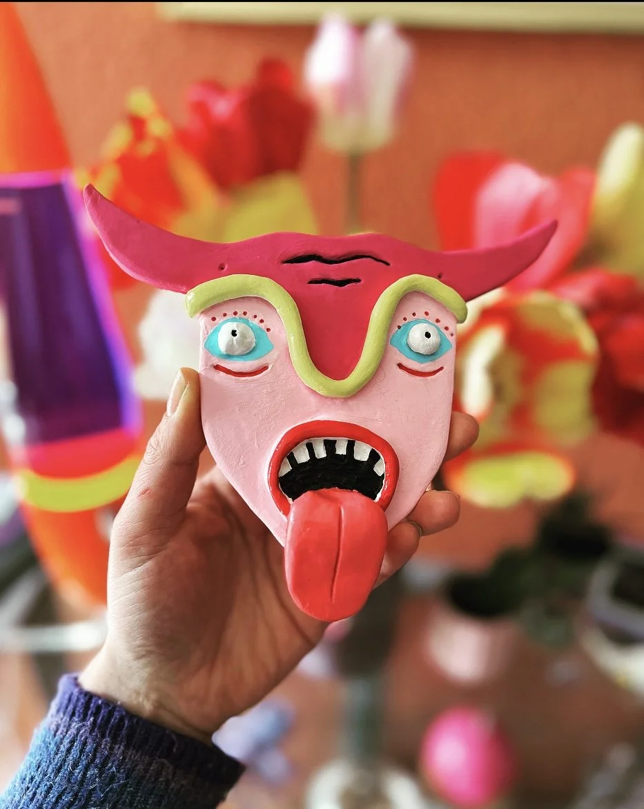 A colorful ceramic sculpture of a face with exaggerated features, including blue eyes, a red tongue sticking out, and a pink face, held in a person's hand with a blurred background of various colorful sculptures and flowers.