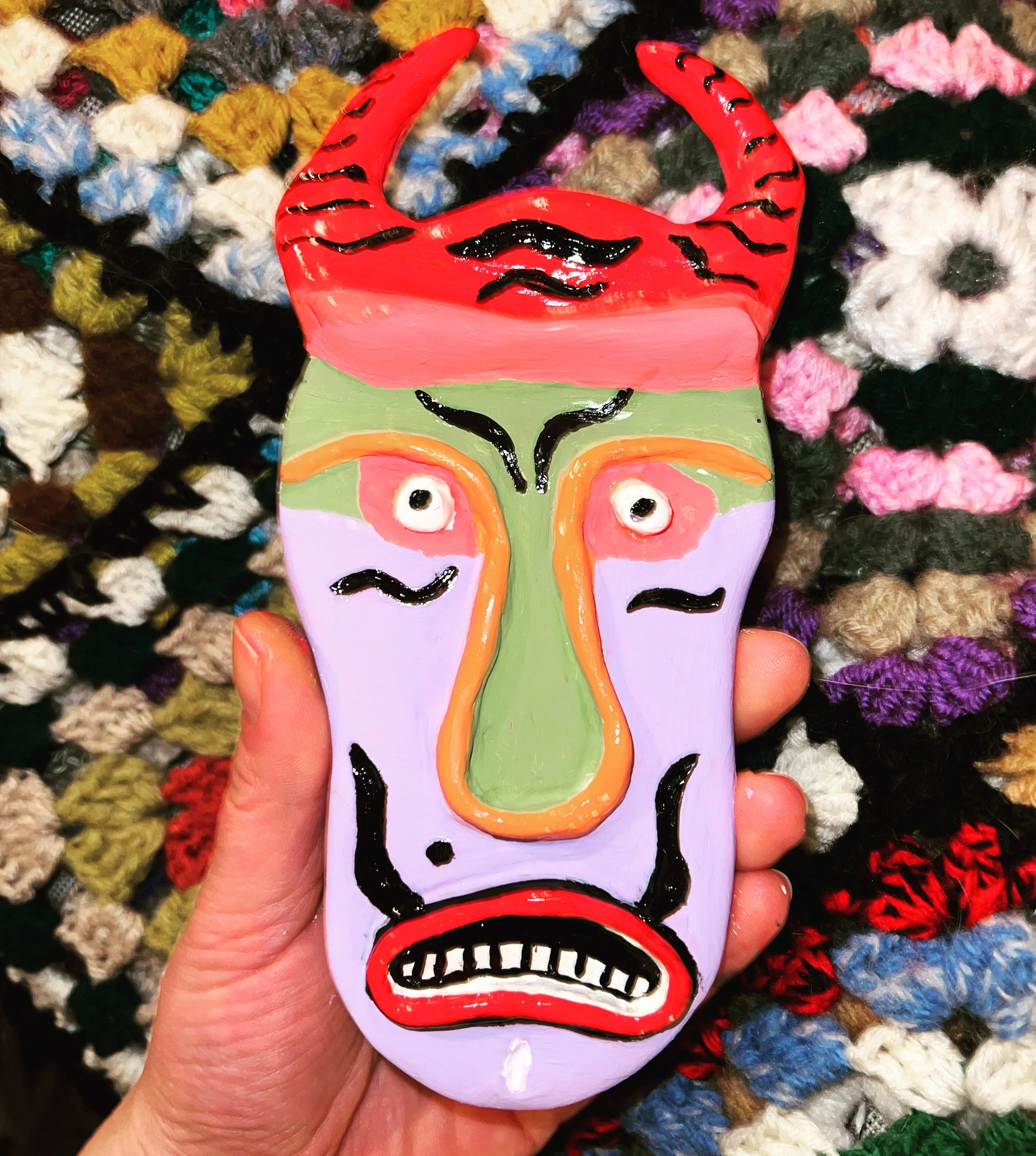 Colorful, abstract face mask with exaggerated facial features, including wide eyes, a long green nose, and an open mouth, being held against a multicolored woven fabric background.