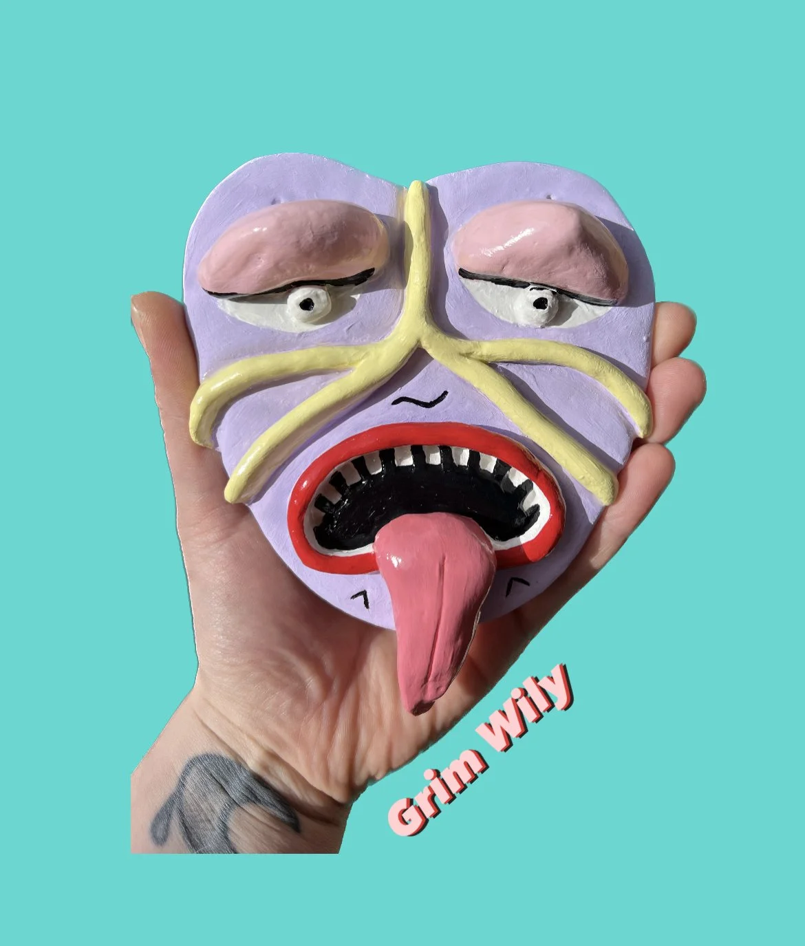 A person holds a colorful, artistic mask with a exaggerated, grimacing face and tongue out against a turquoise background. The mask has purple skin, closed eyes with pink eyelids, yellow lines, a wide black mouth with white teeth and red lips, and a