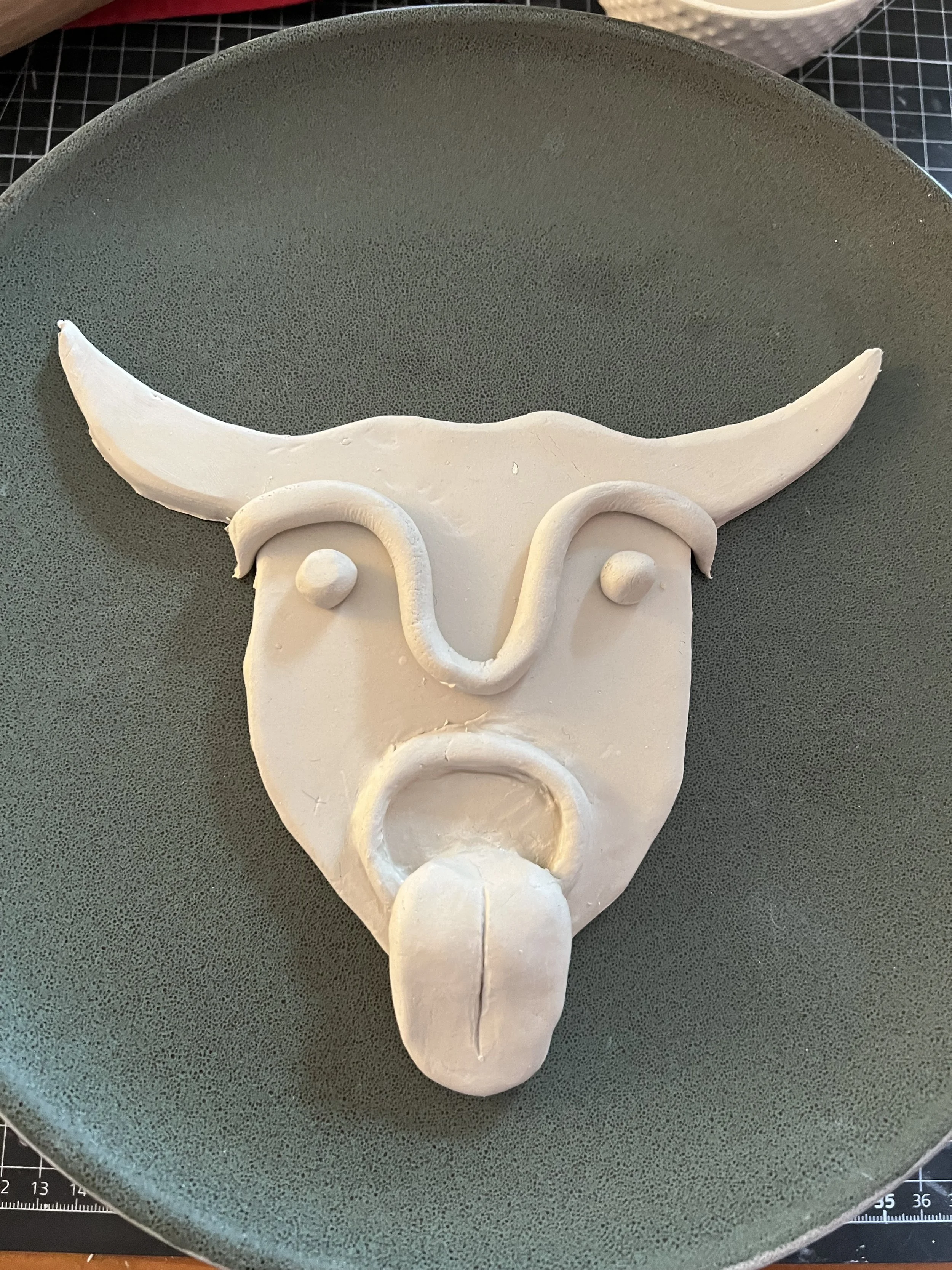Clay sculpture of a bull's face on a green plate, featuring curved horns, outlined eyes, and a protruding tongue.