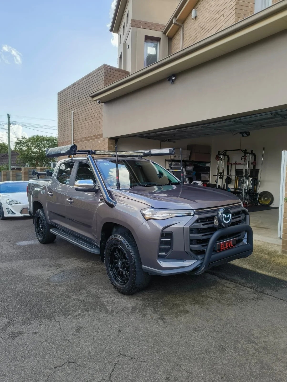 A gray pickup truck parked in front of a garage with exercise equipment inside. The truck has off-road tires, a bull bar on the front, and equipment mounted on the roof, including a light bar, a water ski, and other gear.