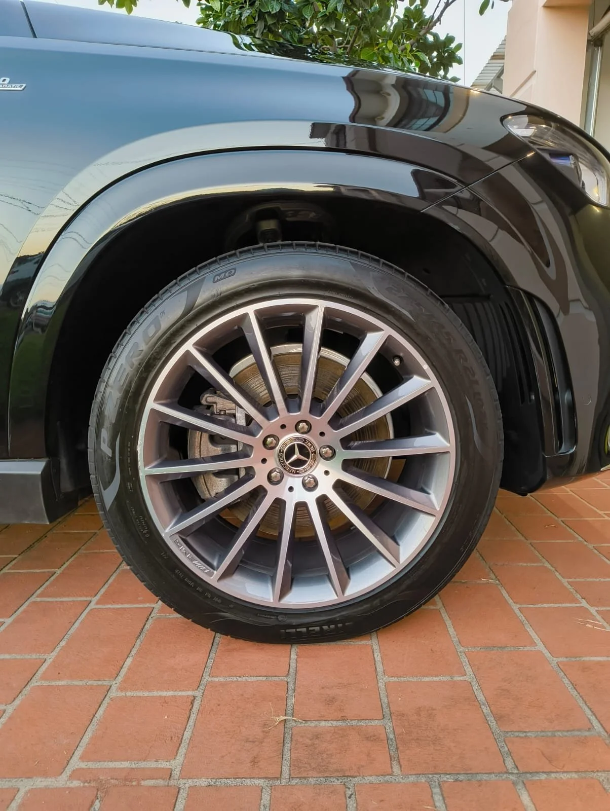 A close-up of a black Mercedes-Benz car's front wheel and tire, parked on a brick driveway, with a tree and a building in the background.
