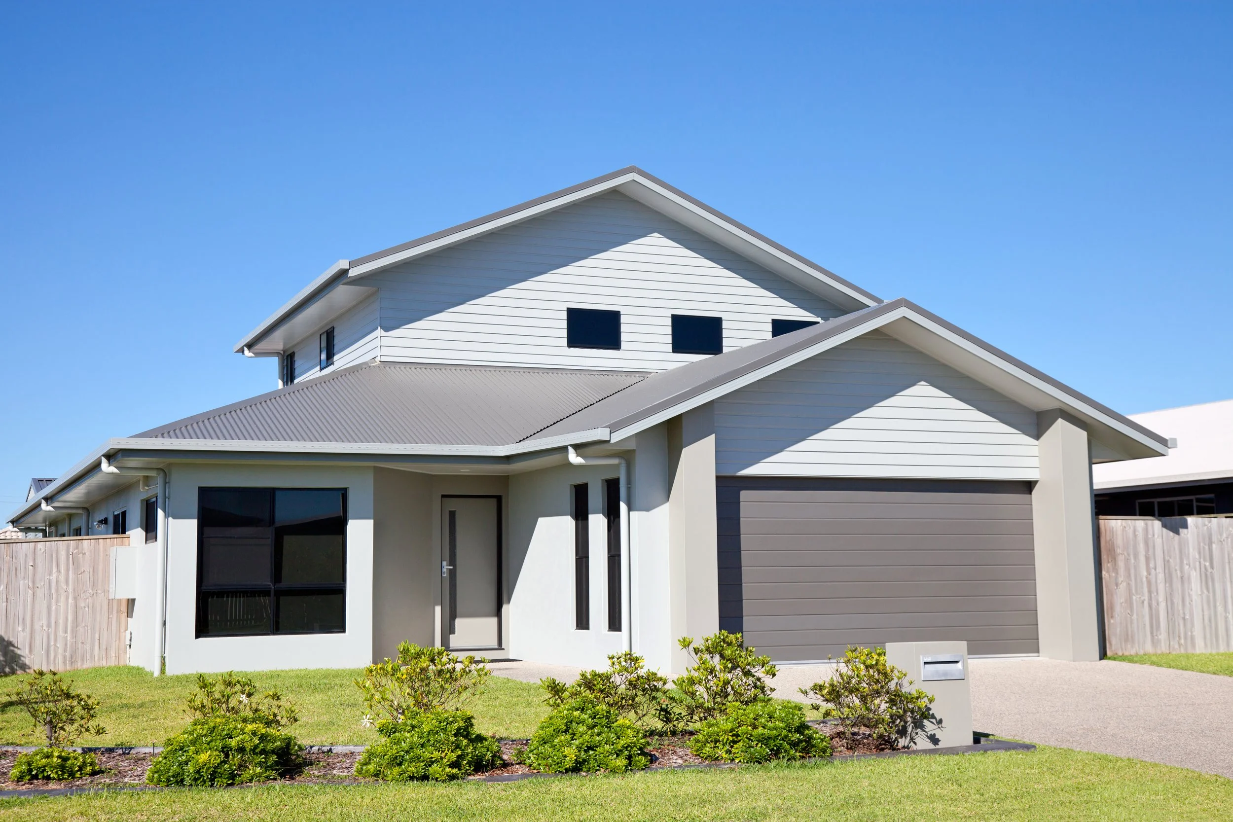 Modern two-story house with a gray garage door, white siding, and a front yard with green shrubs under a clear blue sky.