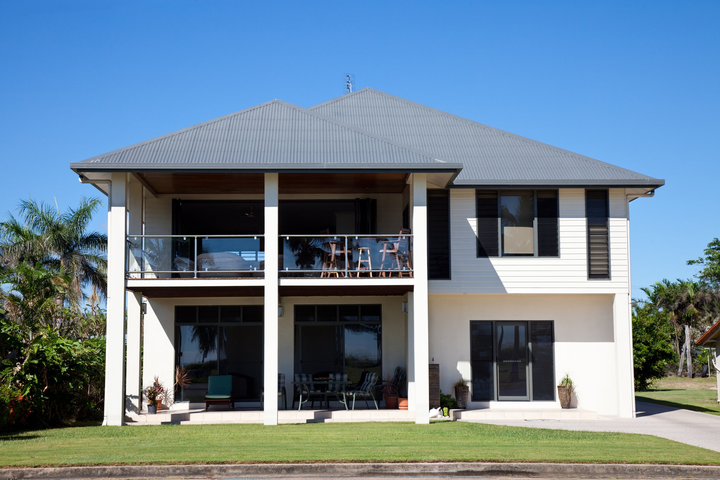 Two-story white house with black window frames, a gray metal roof, and a balcony with outdoor seating. The house is surrounded by green grass, palm trees, and a clear blue sky.