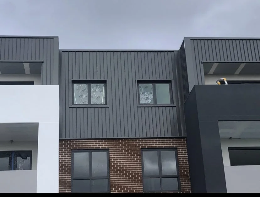 Close-up of a modern multi-story building with gray metal siding, red brick, and glass windows under a cloudy sky.