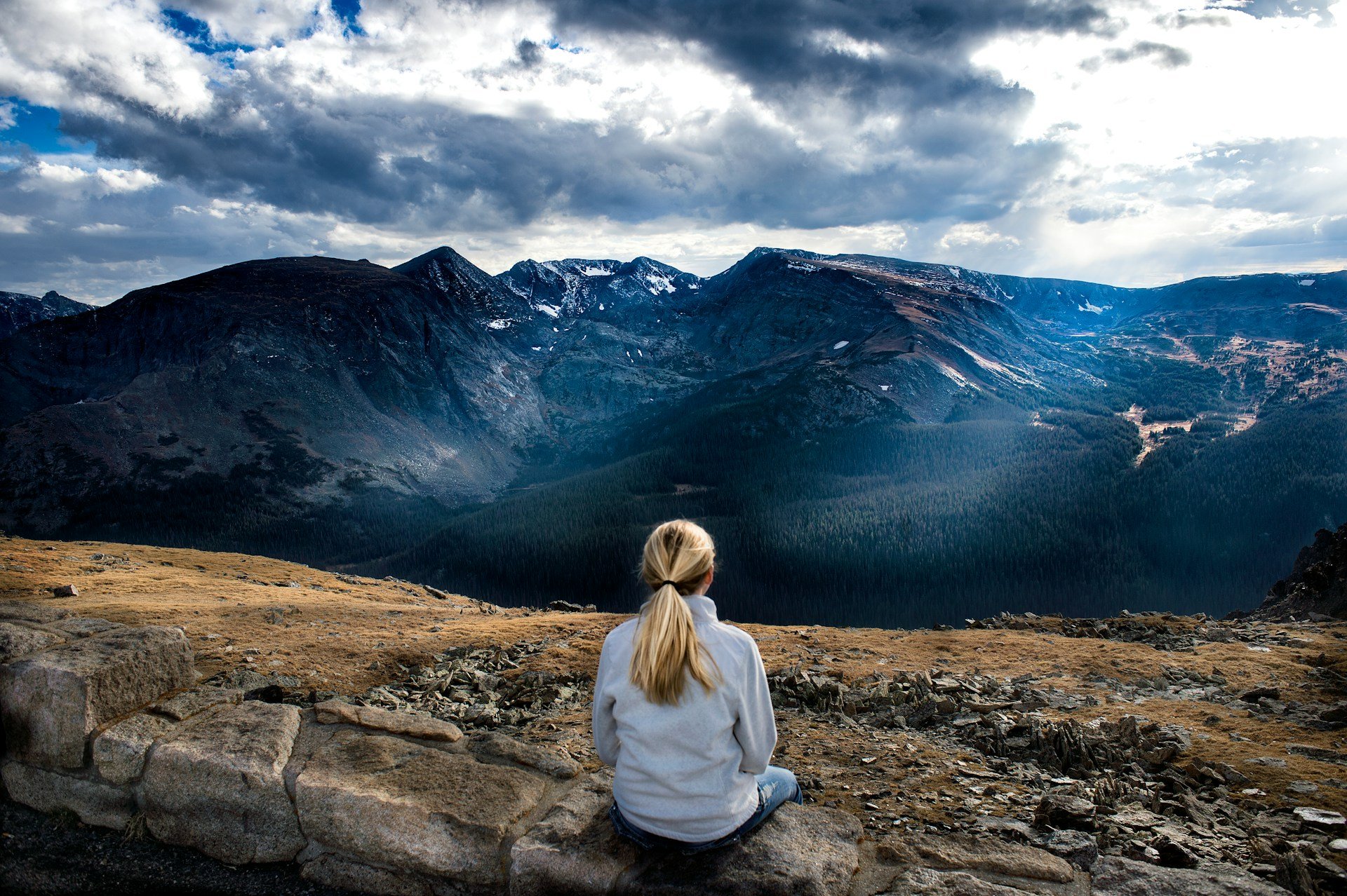 Eine Frau sitzt auf Steinen in einer bergigen Landschaft, blickt auf die Berge und Wolken am Himmel, während sie eine weiße Jacke trägt und ihre Haare zu einem Pferdeschwanz gebunden sind.