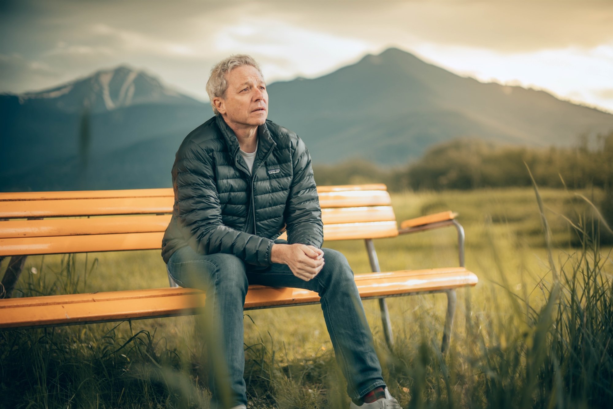 Ein älterer Mann sitzt nachdenklich auf einer Holzbank in einer Naturlandschaft mit Bergen im Hintergrund. Es ist bewölkt, und das Licht ist sanft.