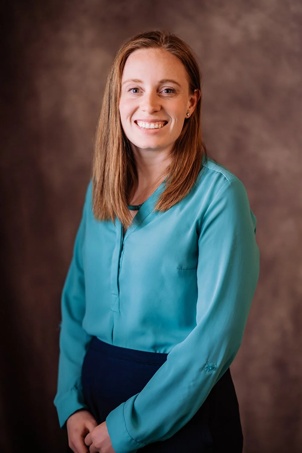 Woman in a teal blouse smiling in a portrait photo with a brown background.
