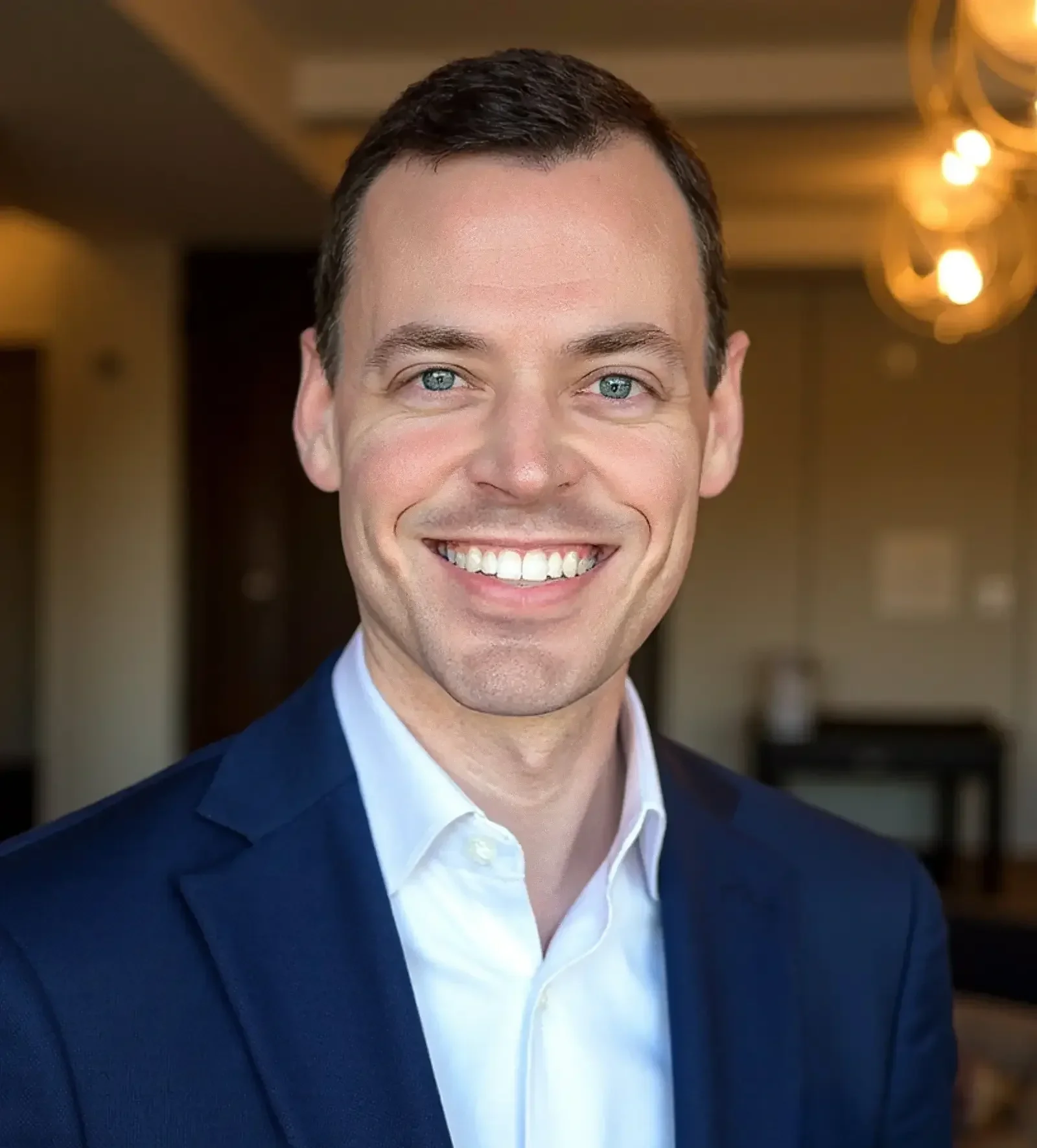 Patrick Gregoire wearing a navy blue suit and white shirt, smiling, indoors with warm lighting and blurred background.