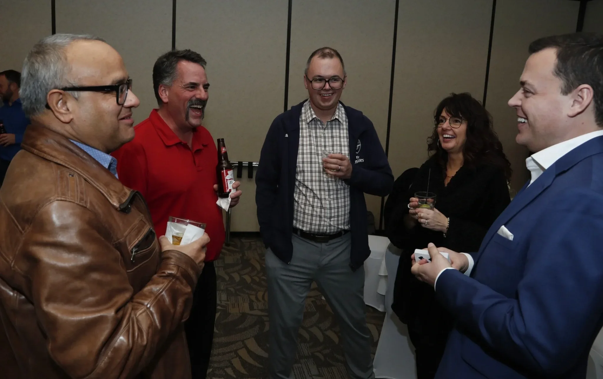 Group of five people, four men and one woman, smiling and talking at a social gathering indoors, holding drinks.