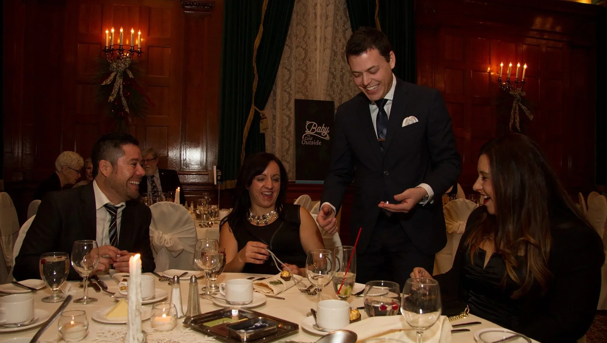 People at a formal dinner table joyful and smiling, with Winnipeg magician Patrick Gregoire entertaining them with close-up magic at a corporate holiday party.