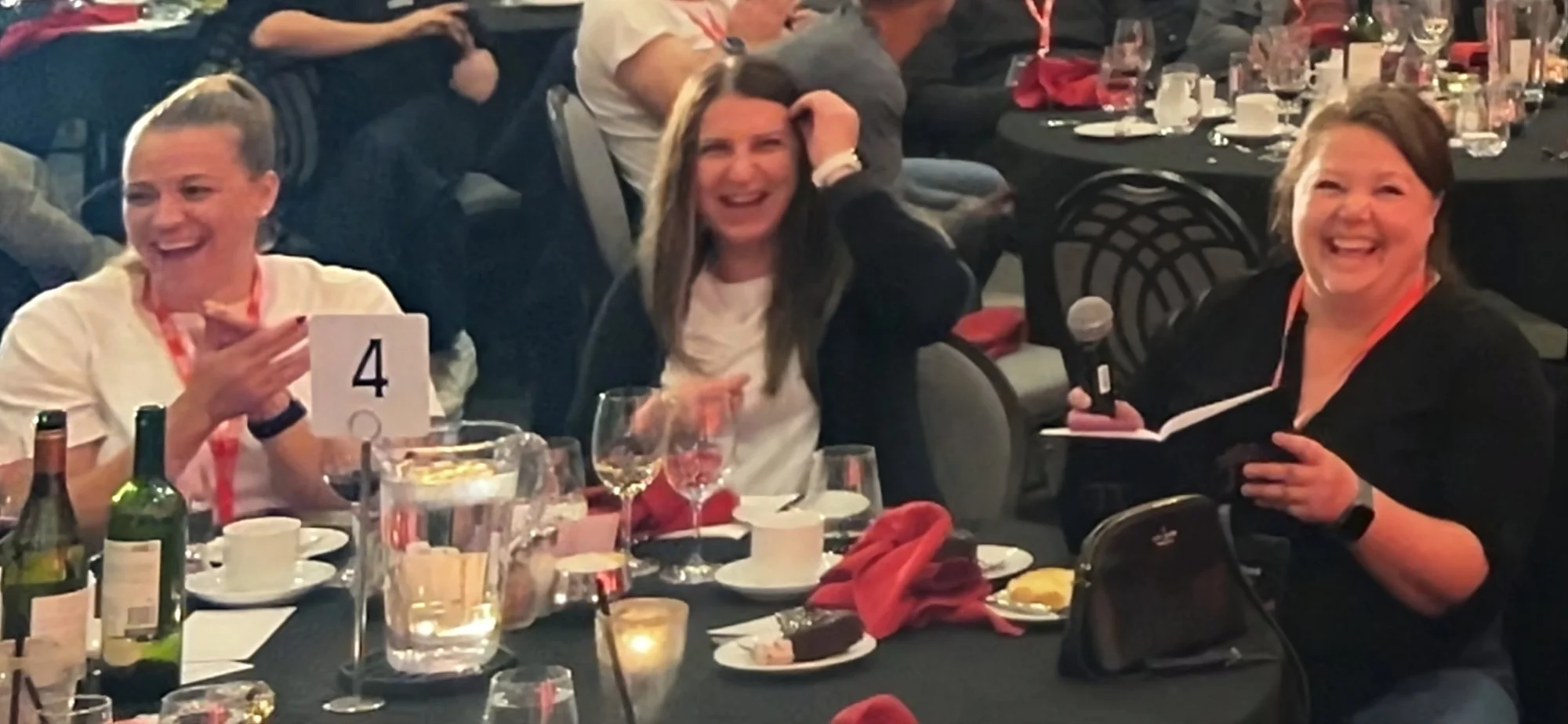 People laughing and enjoying a meal at a banquet table with glasses, bottles, and plates, during a social event.