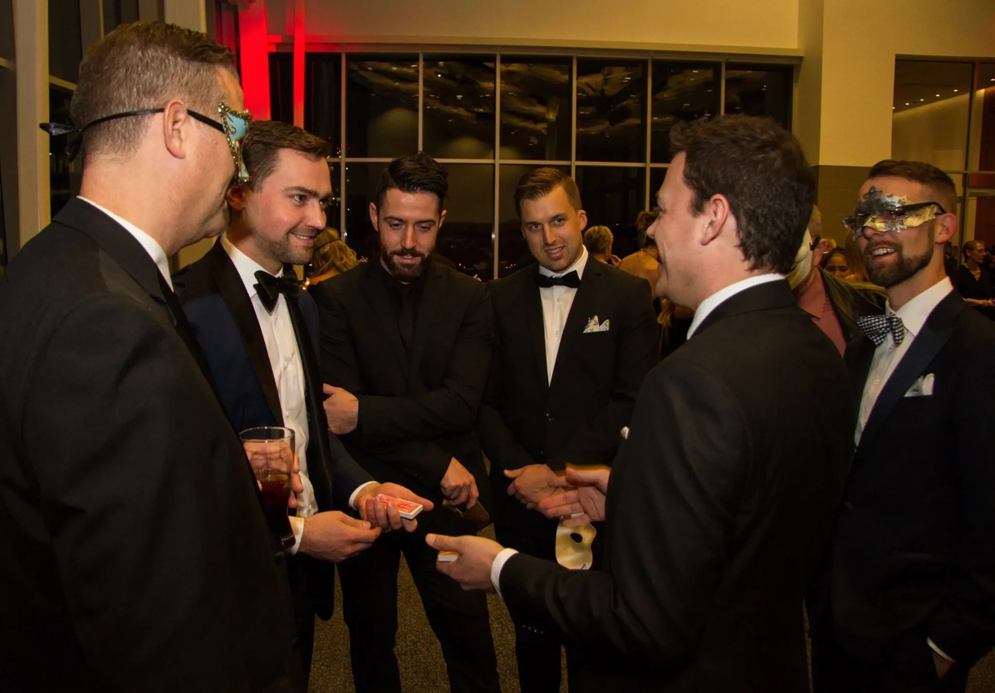 Group of men in tuxedos and masks enjoying some close-up magic with Patrick Gregoire at a formal event.